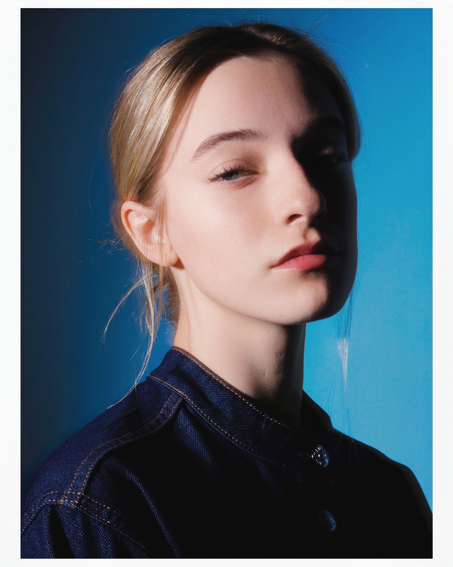 A portrait against a vibrant blue background showcases contemplative pose in denim attire with blonde hair swept back.
