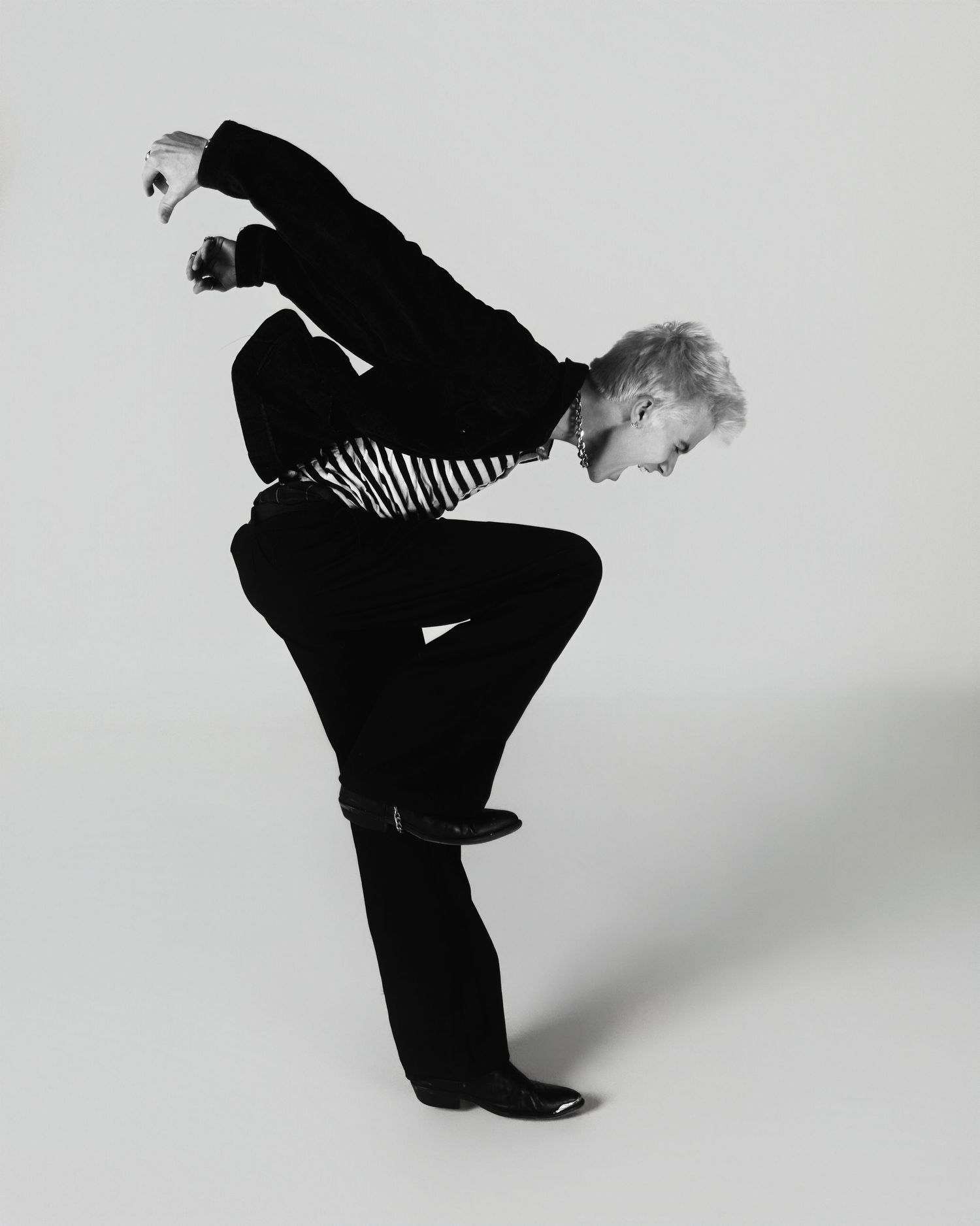 A person in a black outfit with a striped shirt strikes a dramatic pose against a white backdrop in this monochrome photo.