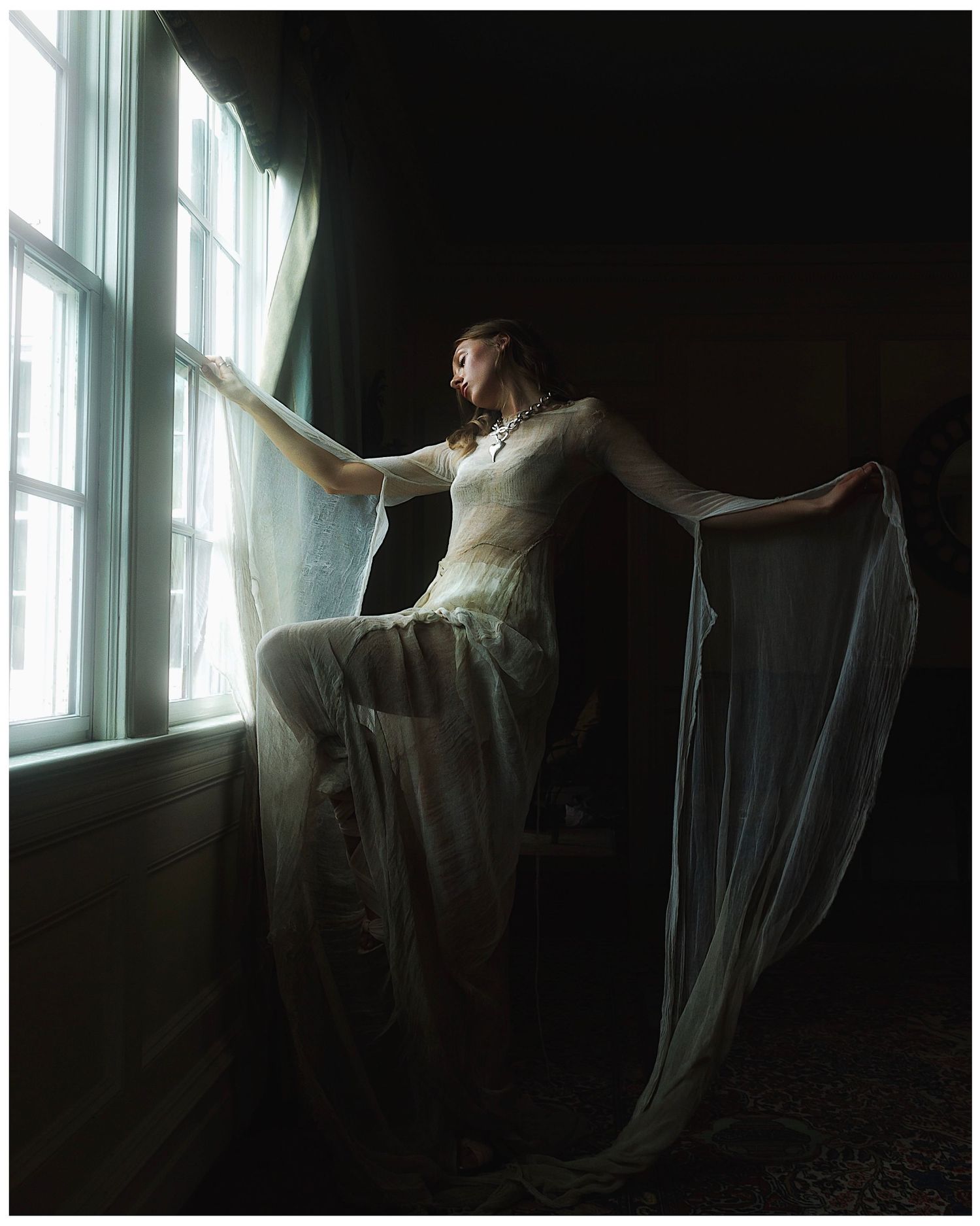 A woman in a flowing white gown stands by a bright window in a dark room, gracefully spreading her sheer veil wide.