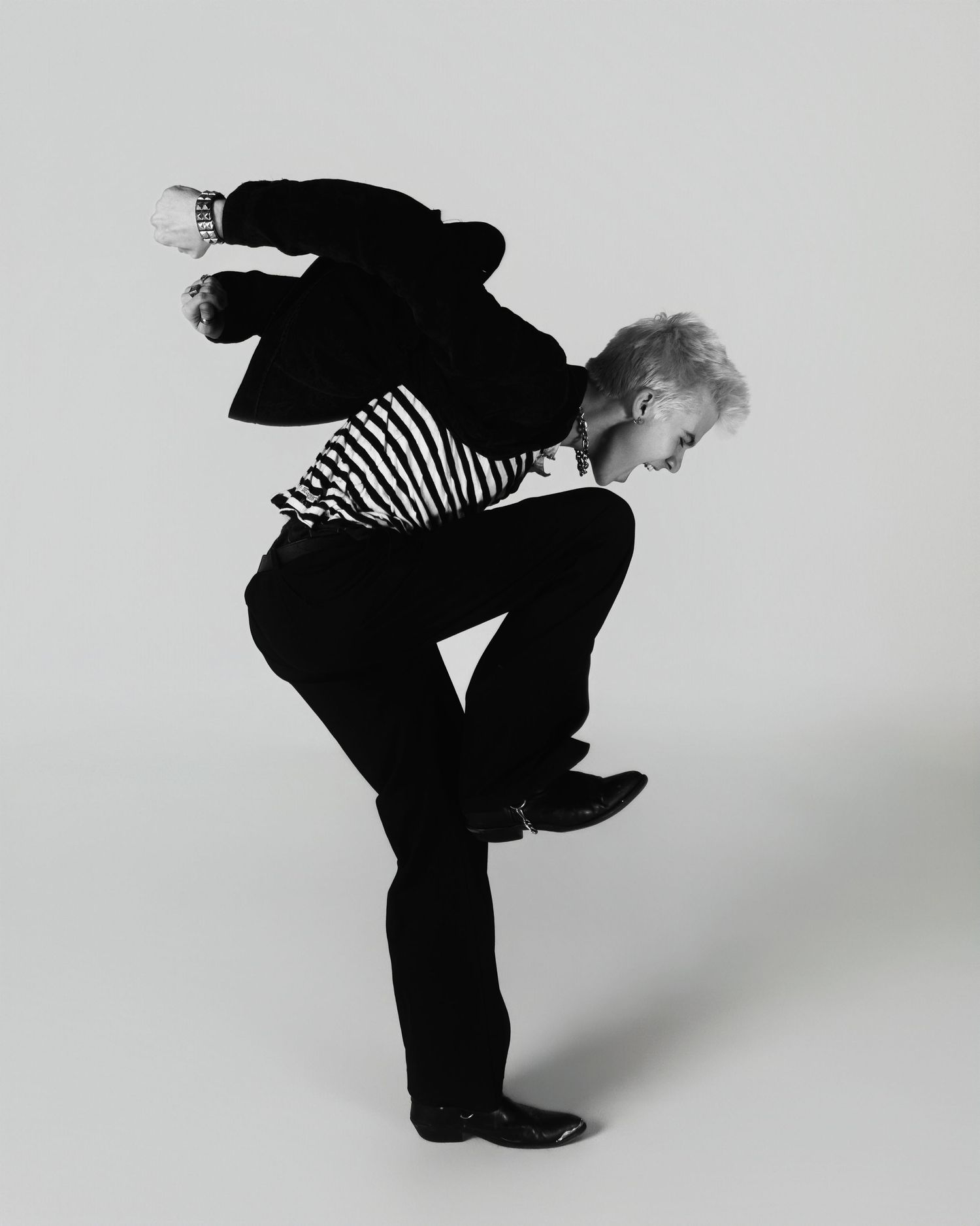Black and white artistic photo of a person in striped top and black pants jumping in dynamic bent-over pose against plain backdrop.