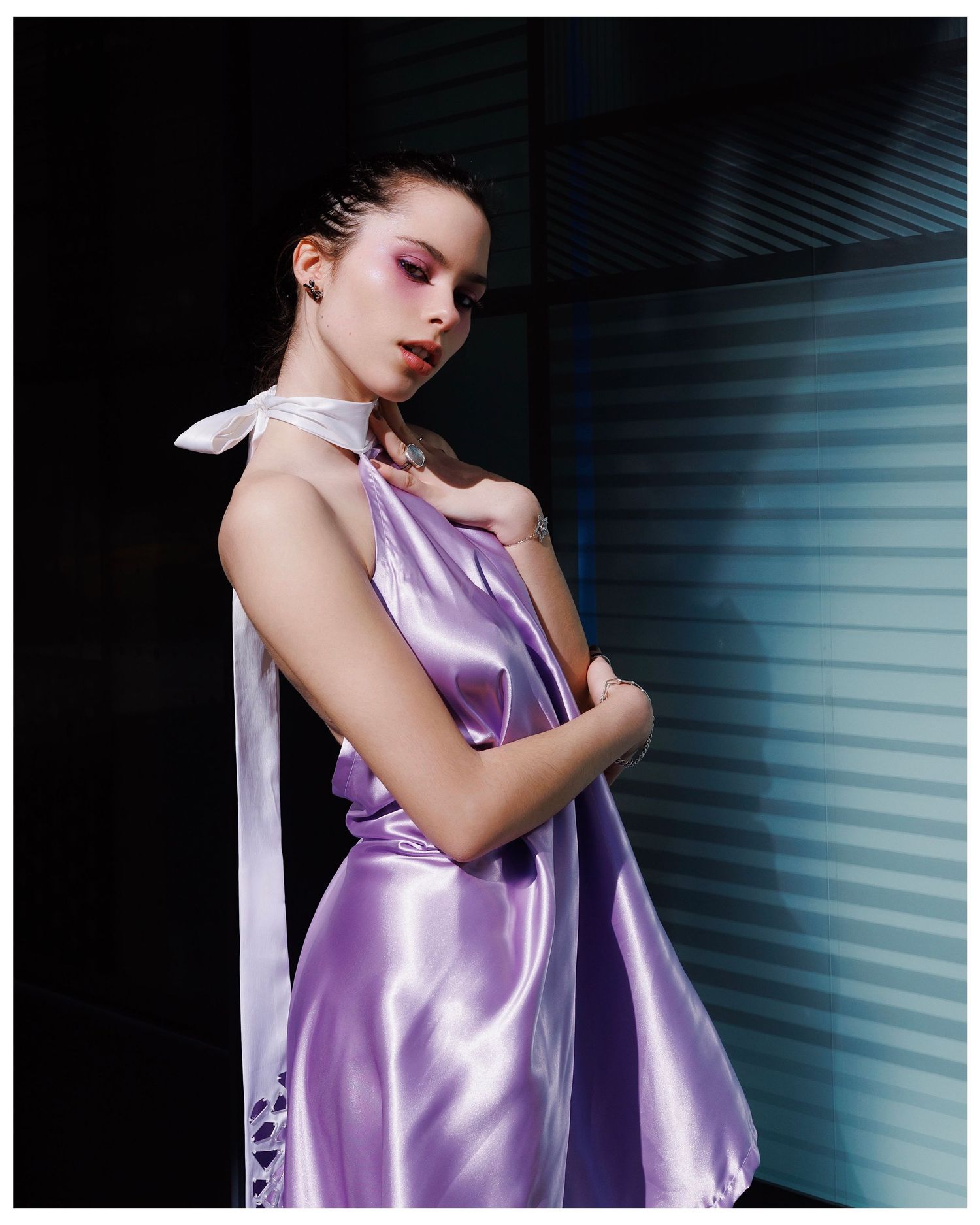 Figure in shimmering lilac silk dress poses against reflective window blinds creating dramatic lighting effects.