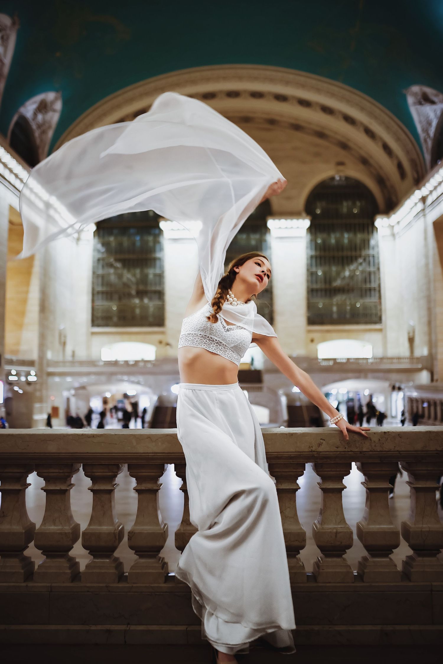 A person in an elegant white dress strikes graceful poses by a classic marble balustrade in a grand arched hall.