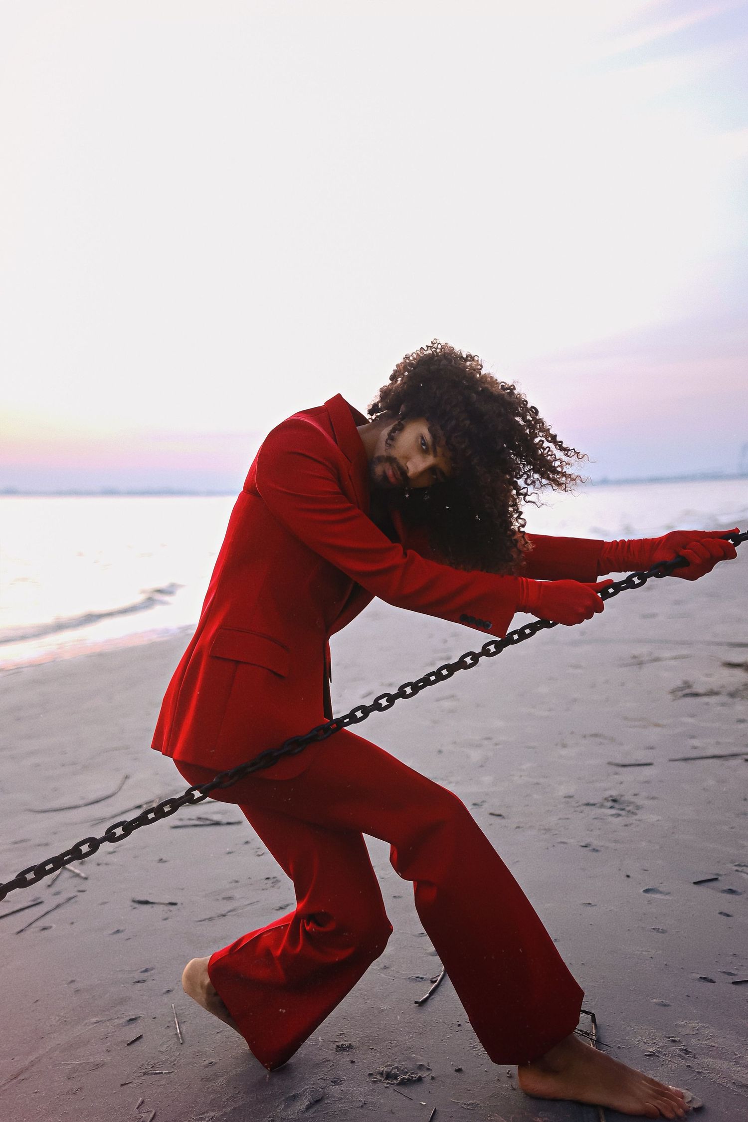A dancer in a red outfit performs dramatic poses while pulling a chain on a beach at sunset.