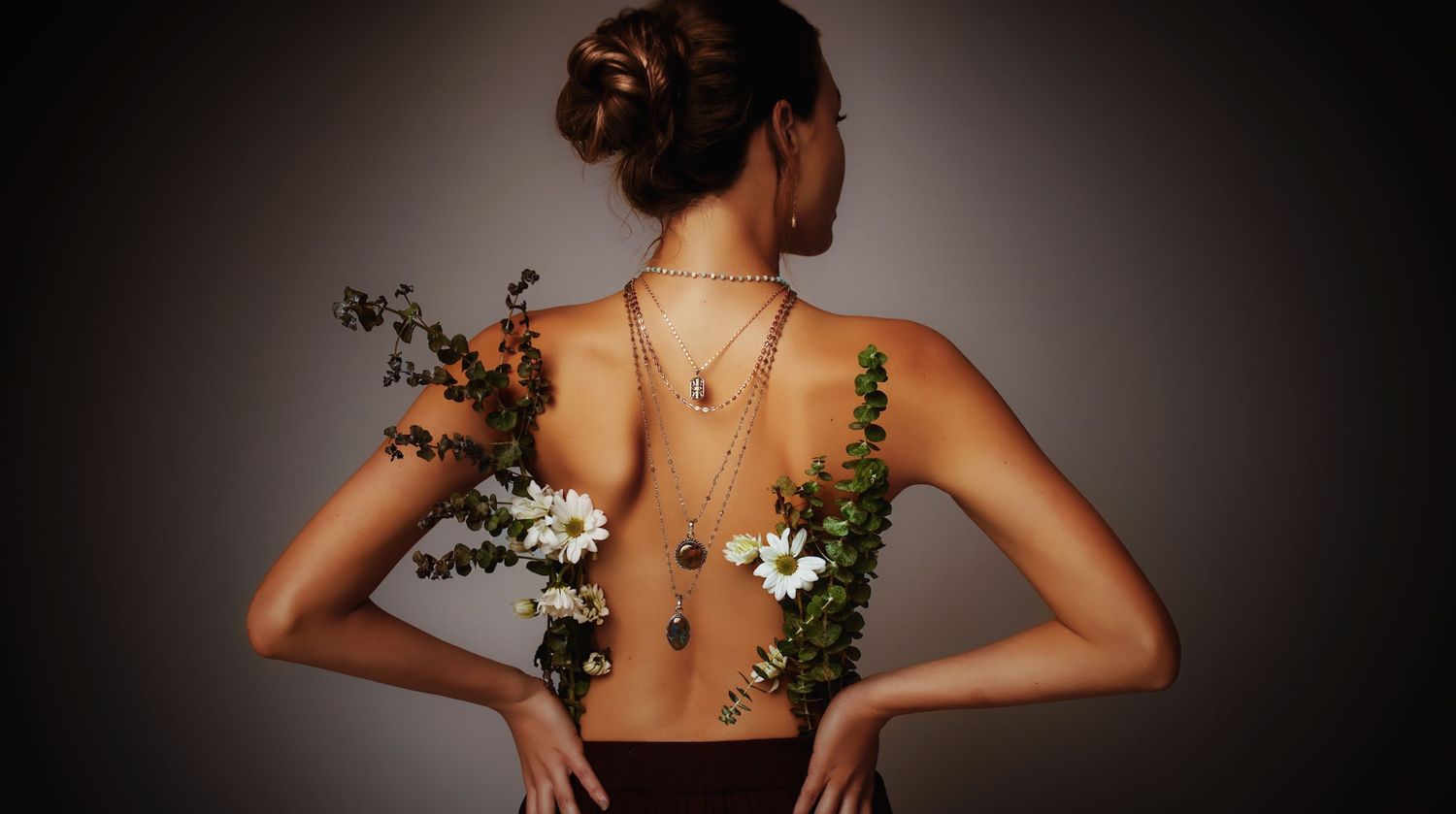 Back view of person with updo hairstyle holding flowers against dark background with delicate neckline jewelry.