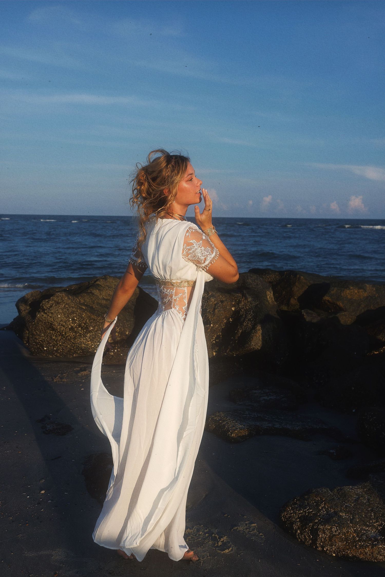 Figure in flowing white dress poses dramatically against rocky coastline and blue ocean at sunset.