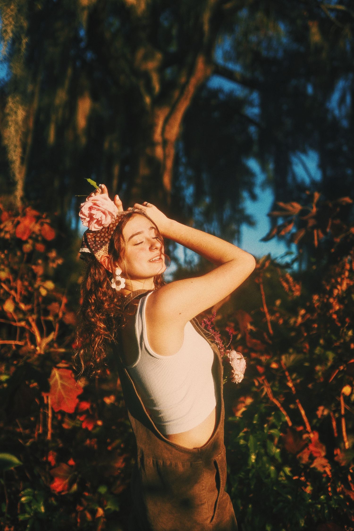 A person in a white tank top dances joyfully at sunset among trees and red foliage in a natural outdoor setting.