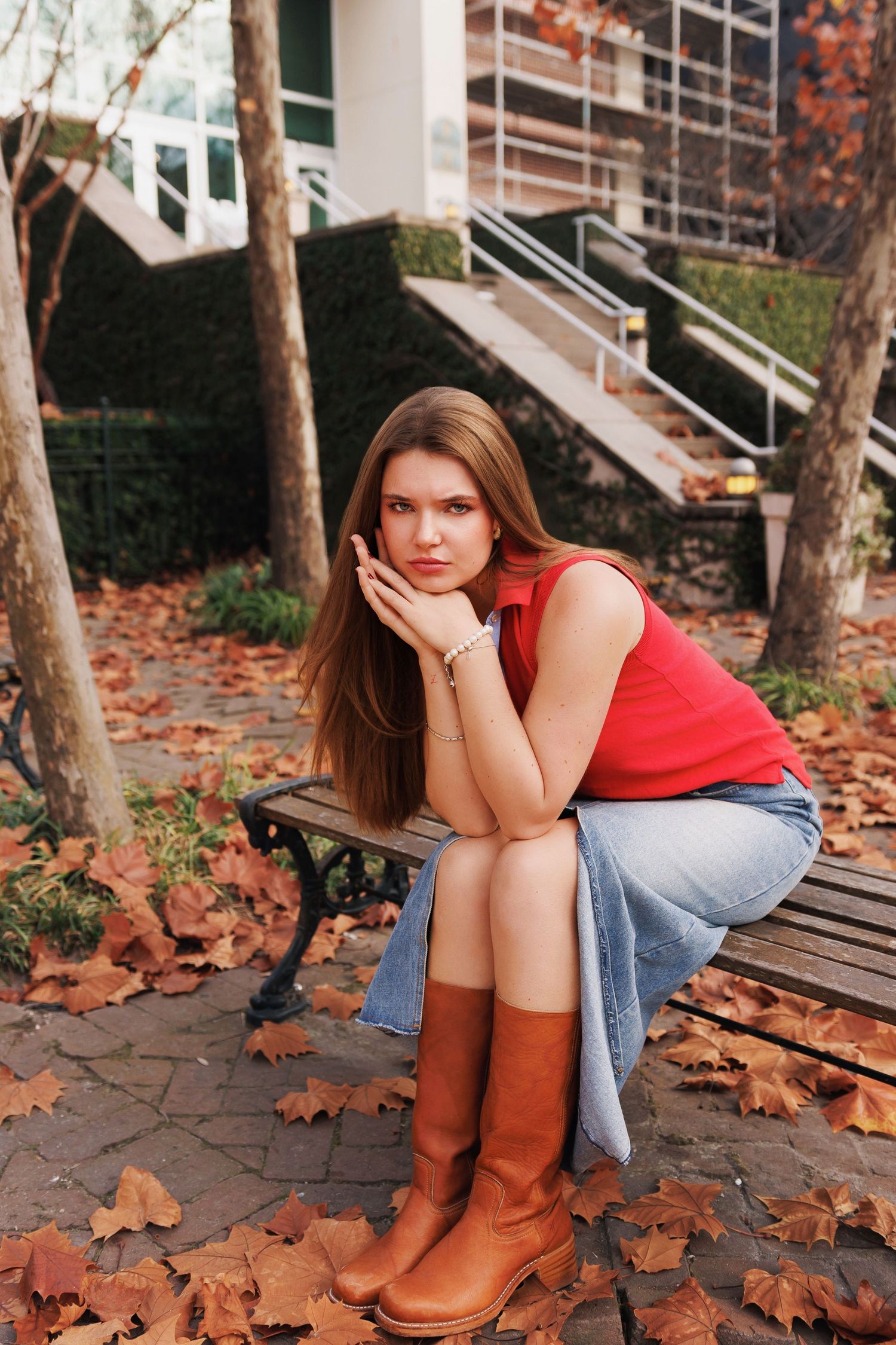 Person in red top and denim skirt sits thoughtfully on outdoor bench surrounded by autumn leaves and brown boots.