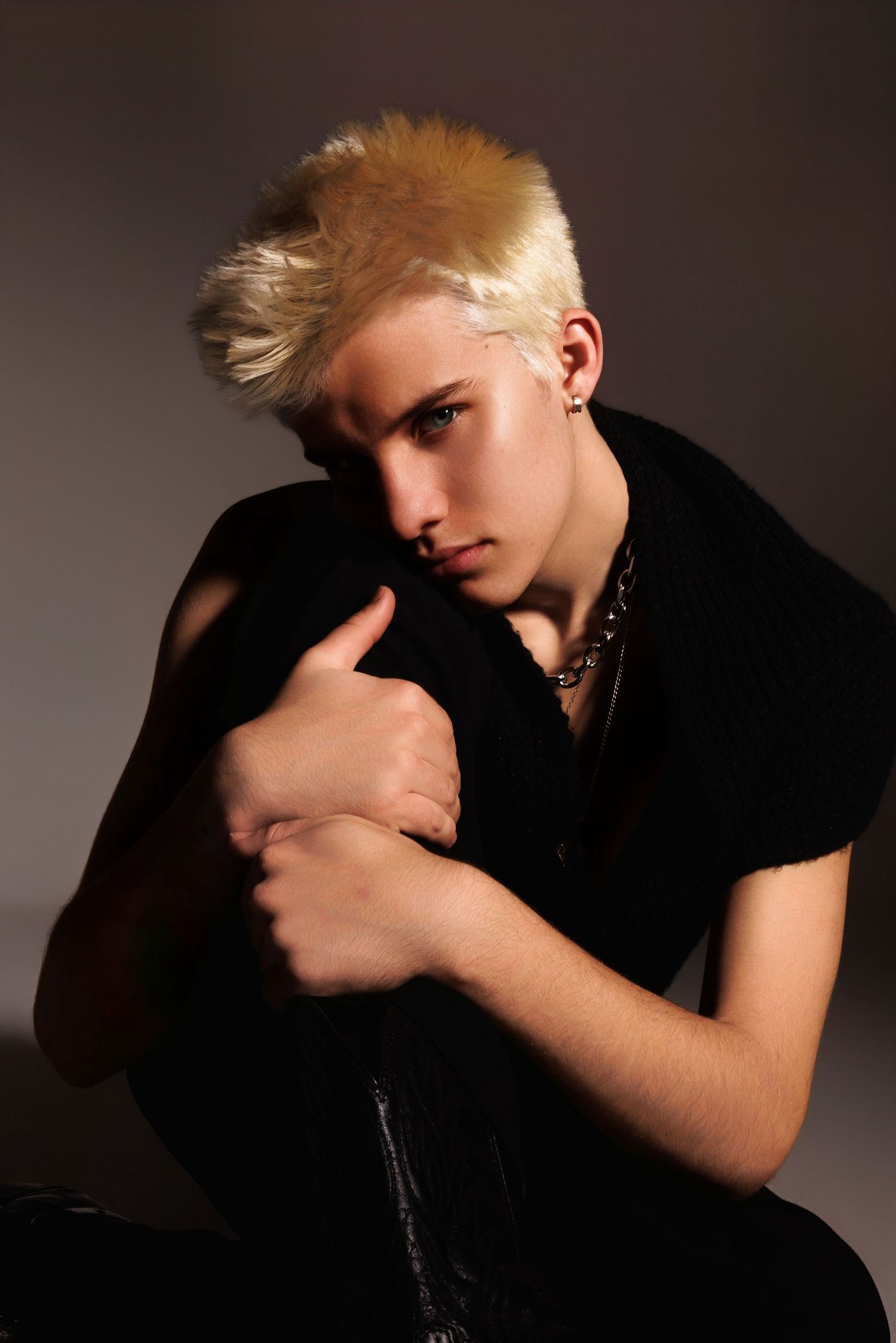 A figure in a black shirt poses against a dark backdrop with dramatic lighting, showcasing blonde spiked hair and jewelry.
