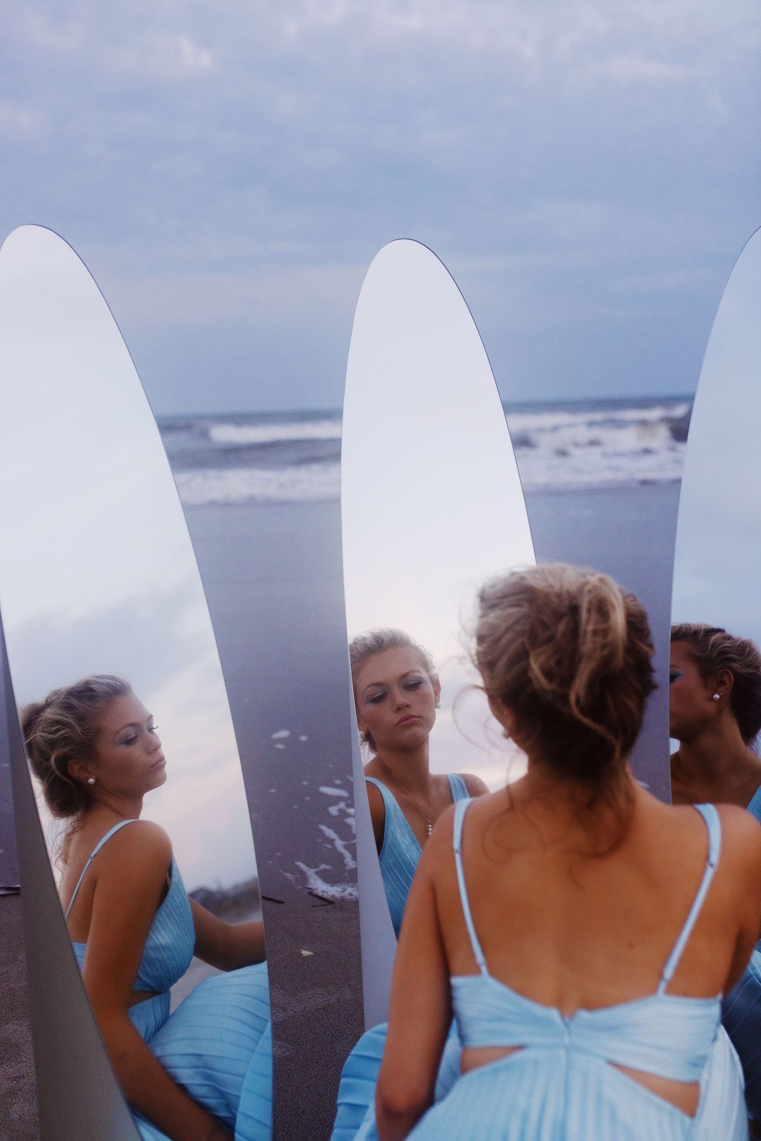 Bridesmaids in light blue dresses stand in a row on a beach holding surfboards at sunset.