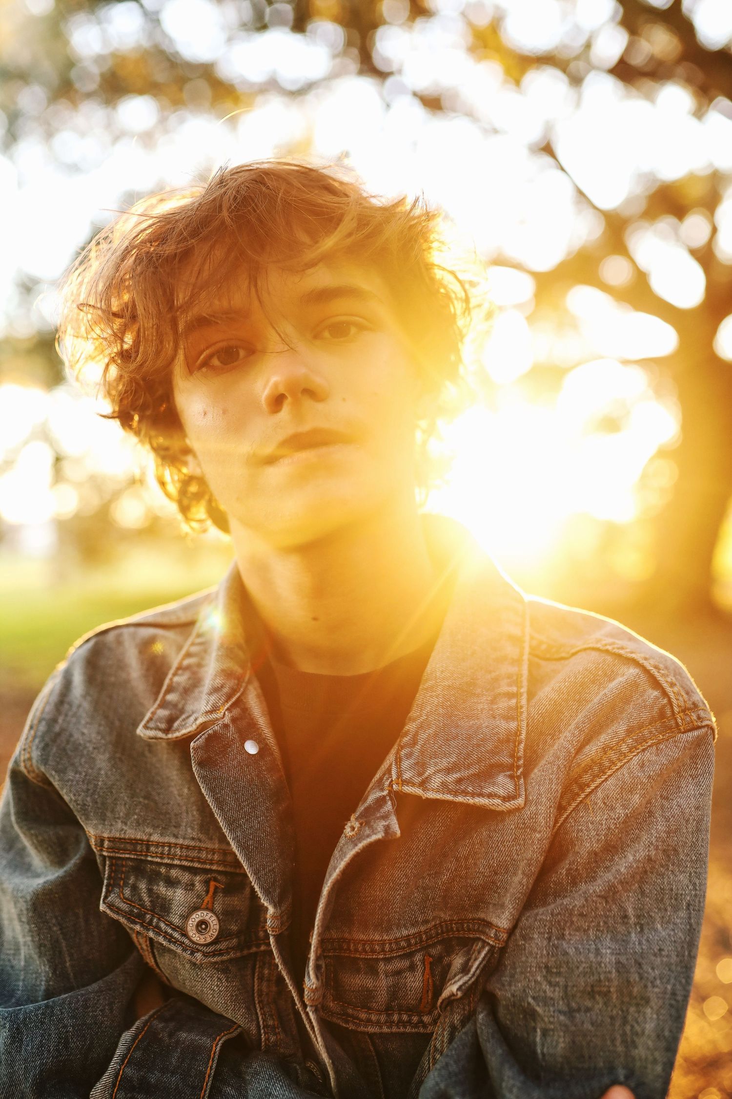 Portrait at golden hour with sunlight streaming through trees behind a denim jacket-clad figure.