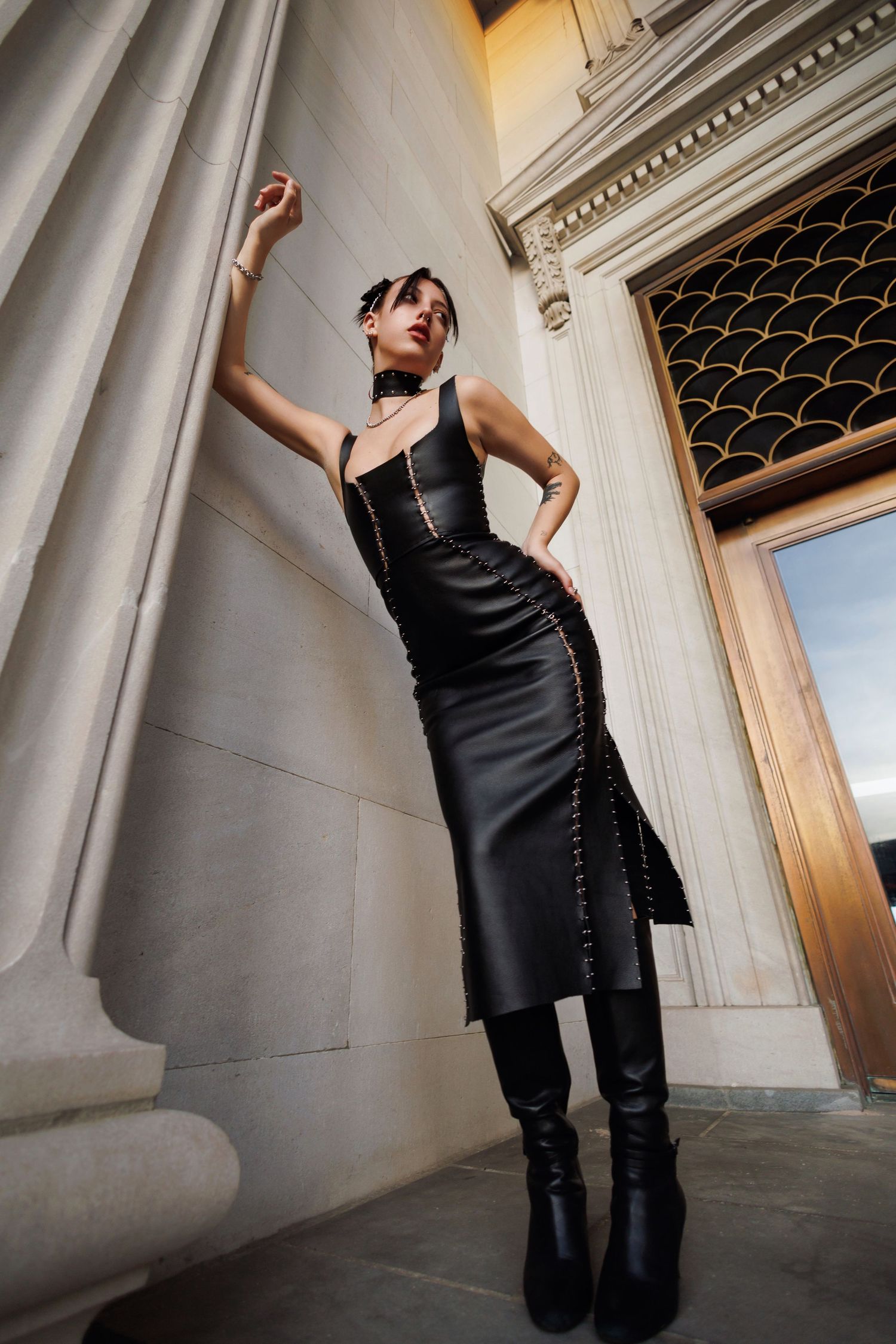 A model in a black leather dress and knee-high boots poses dramatically against classic architectural columns.