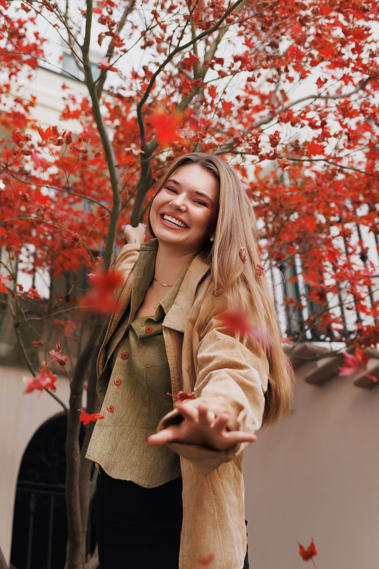 Joyful person in a tan coat enjoying autumn with vibrant red maple leaves falling in the background.