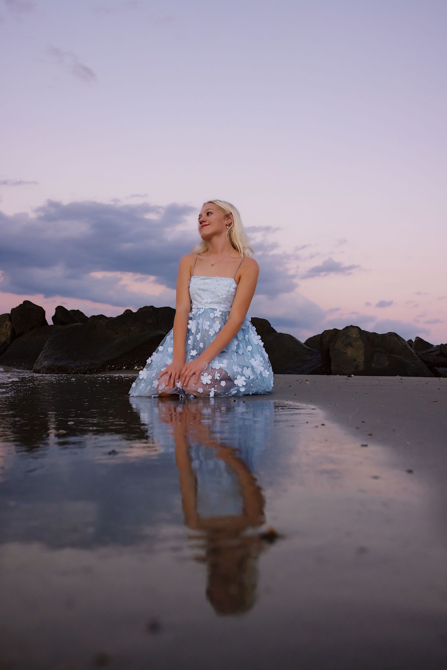 A person in a light blue dress poses on a beach at sunset with dramatic rocky cliffs and reflective wet sand.