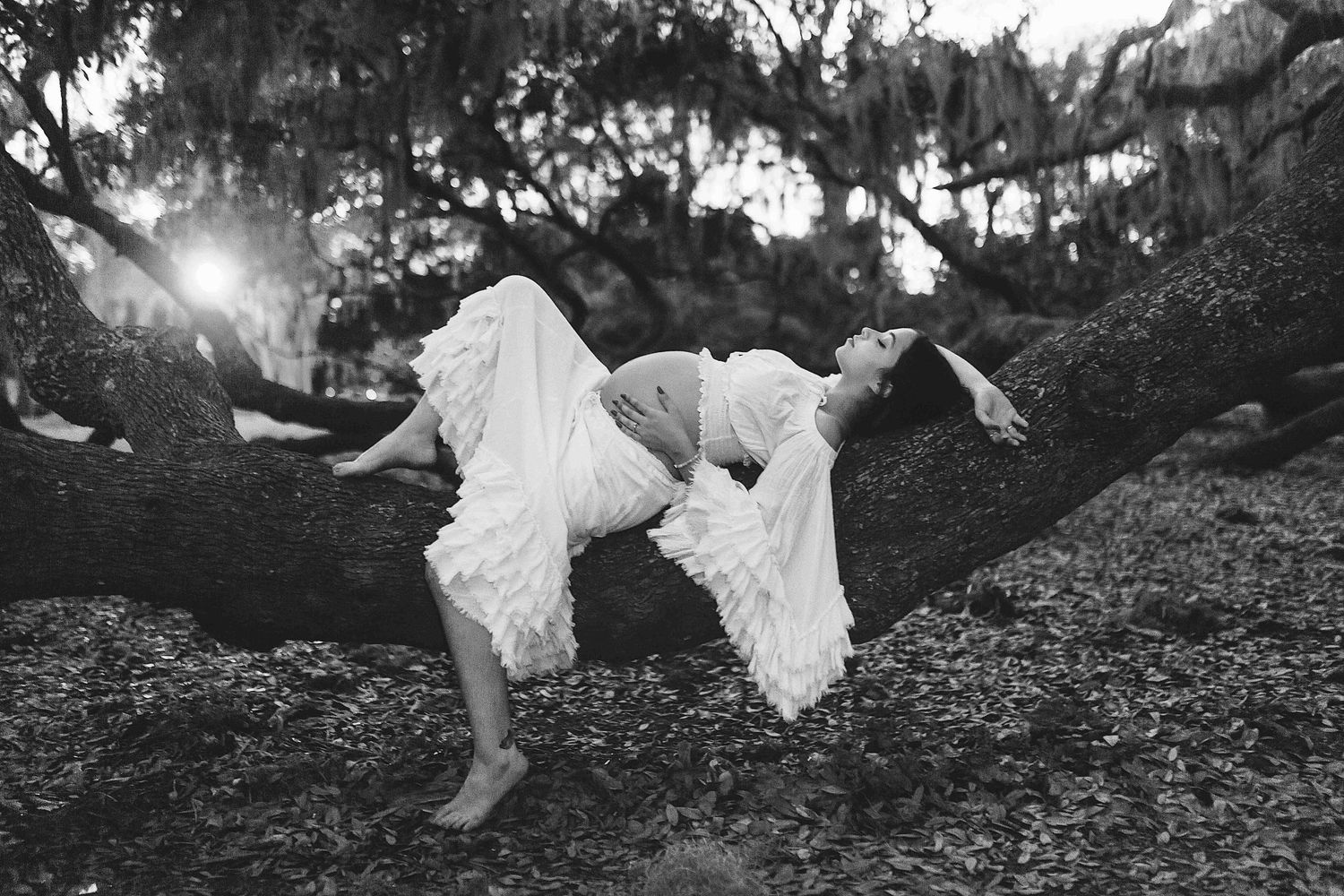 Black and white photo of a woman in a white dress draped over a large tree branch outdoors.