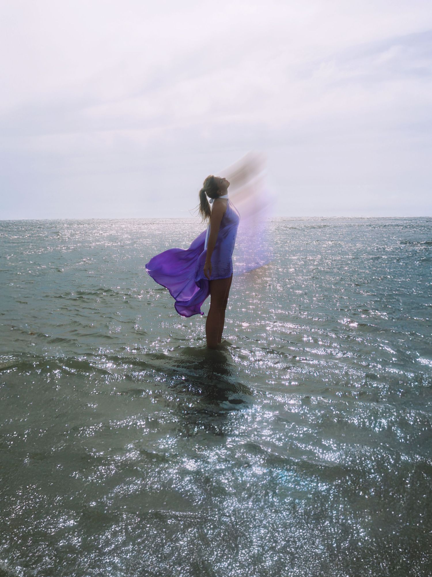 A person in flowing purple fabric stands in shallow ocean waters as sunlight sparkles on the rippling sea surface.