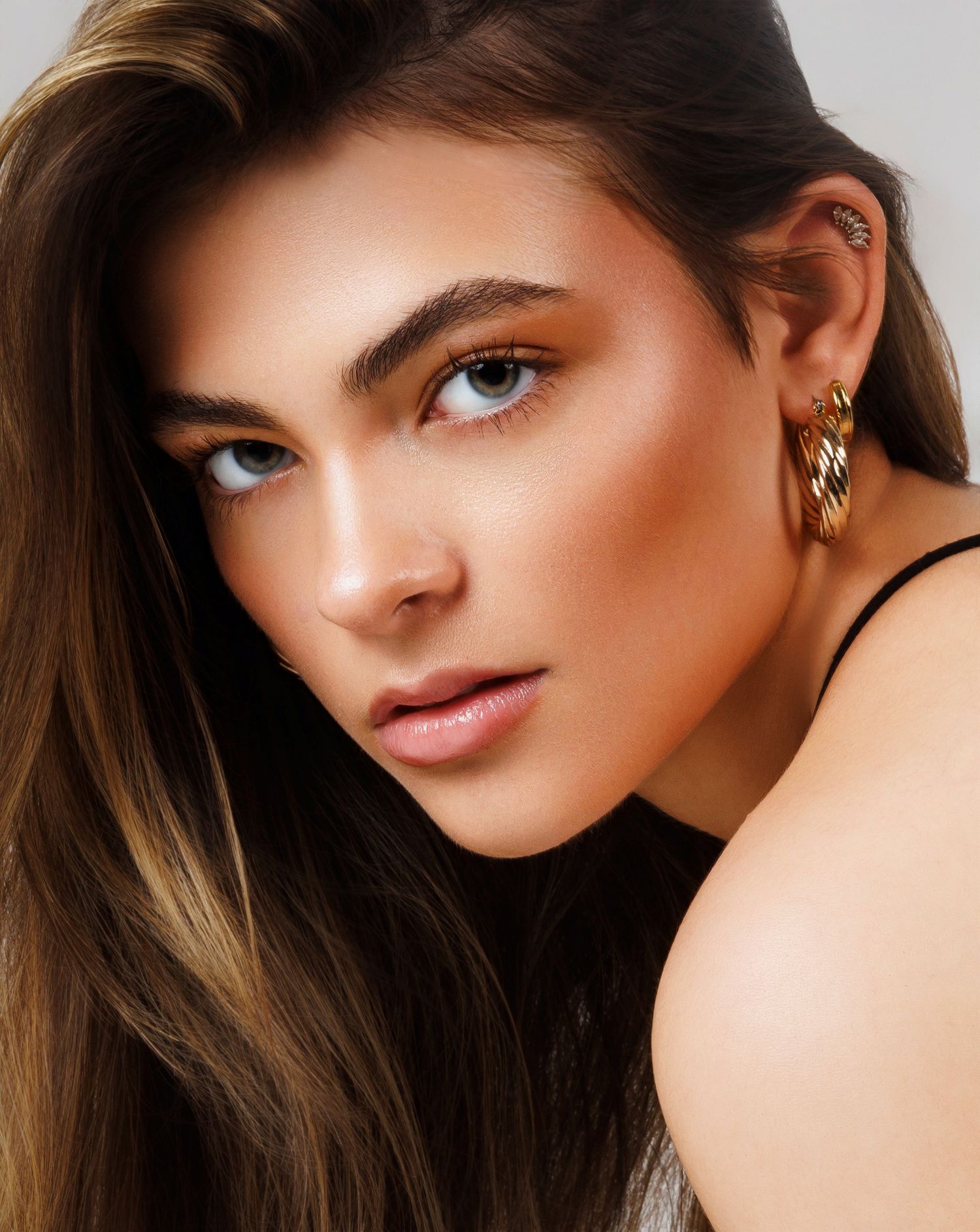 Close-up beauty portrait showcasing luminous bronzed makeup, gold hoop earrings and flowing brown hair.