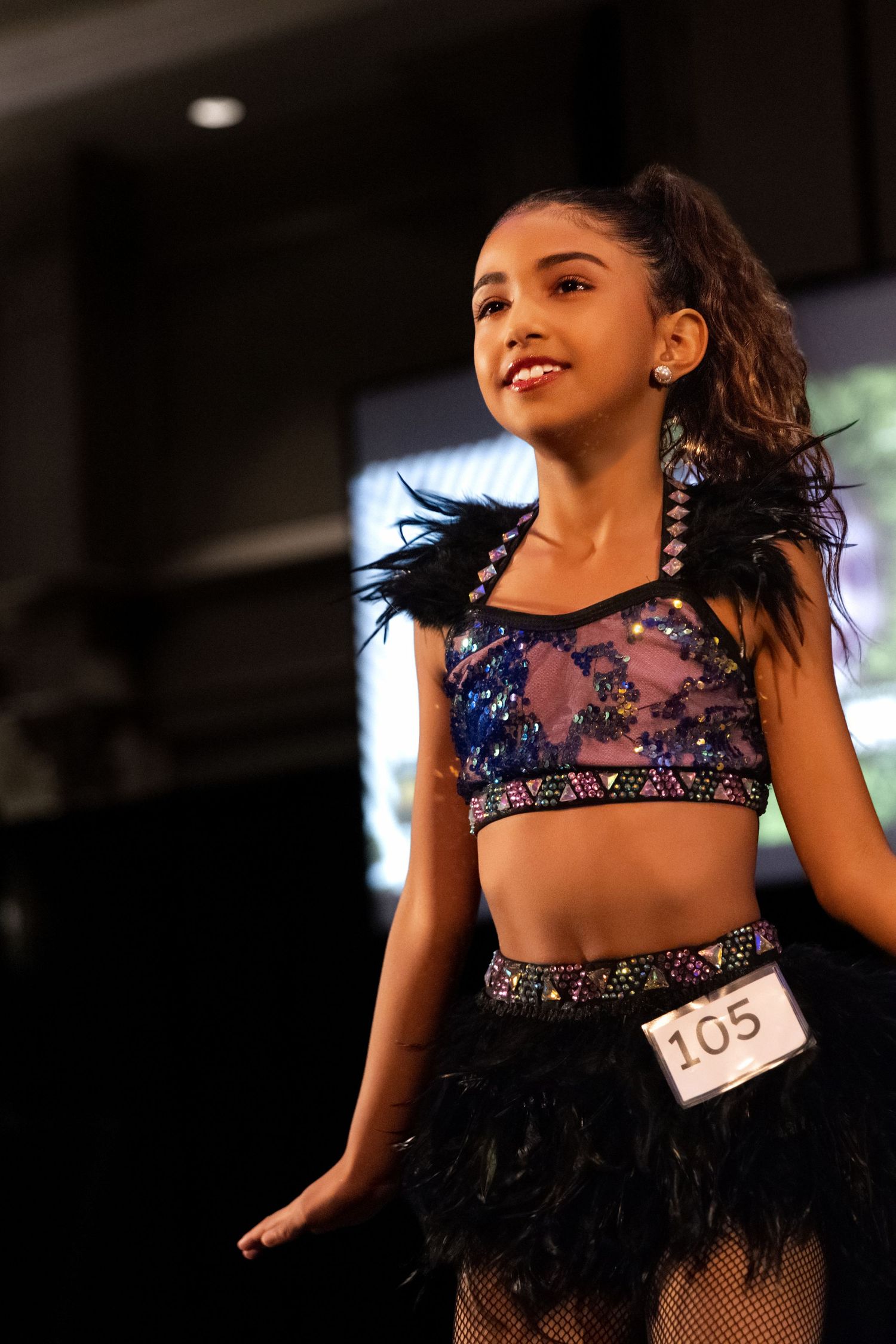 A dancer performs on stage wearing a sparkly blue crop top and feathered black skirt with competition number 105.