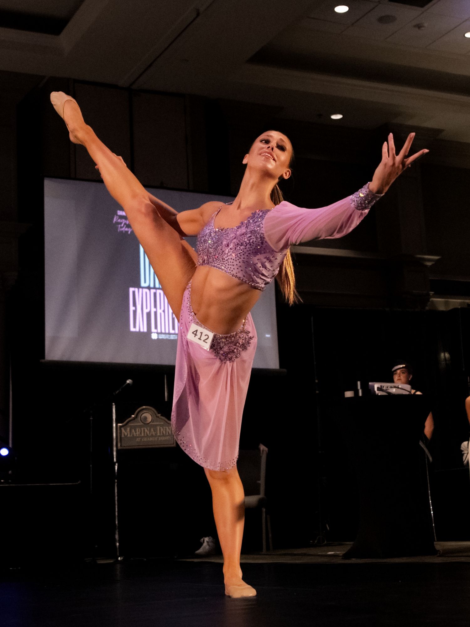 A dancer in pink performs a graceful high kick sequence on a dark stage during a contemporary dance performance.