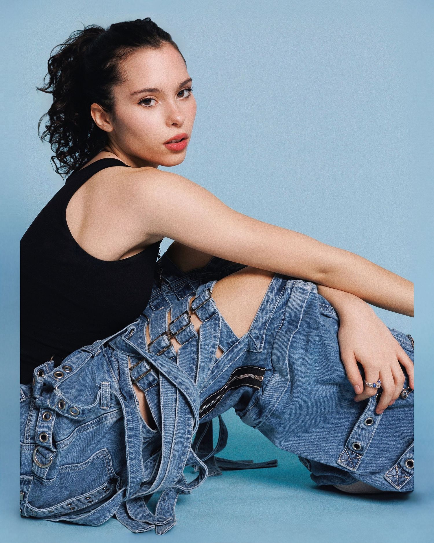 Model wearing black tank top and distressed denim sits against blue backdrop in stylish casual fashion pose.