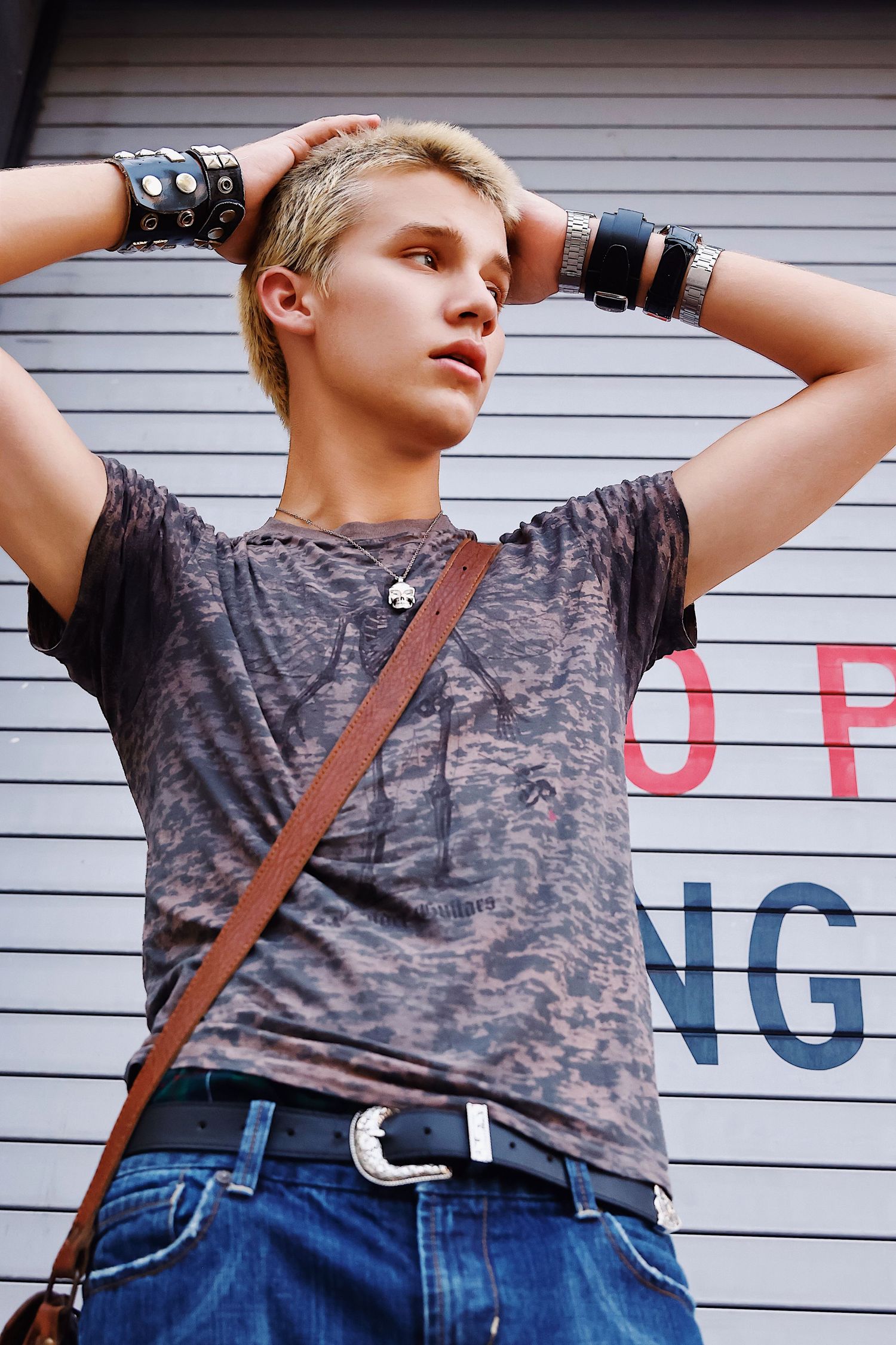 Person in distressed t-shirt and jeans wearing leather wrist cuffs poses against white garage door background.