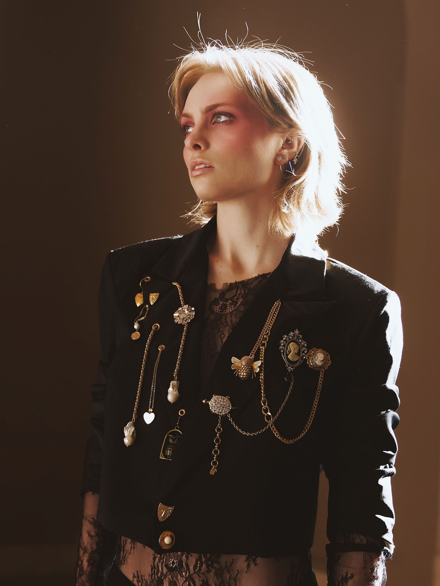 Artistic portrait with dramatic backlighting shows figure in black blazer adorned with vintage brooches and chains.