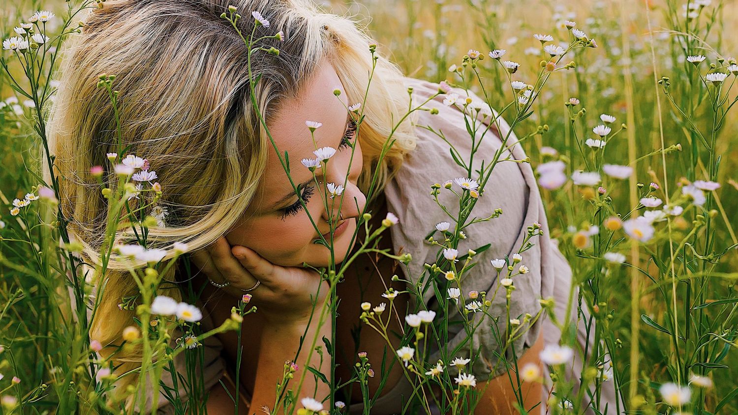 Someone bends down to smell wildflowers in a sunny meadow filled with white daisies and tall grass.