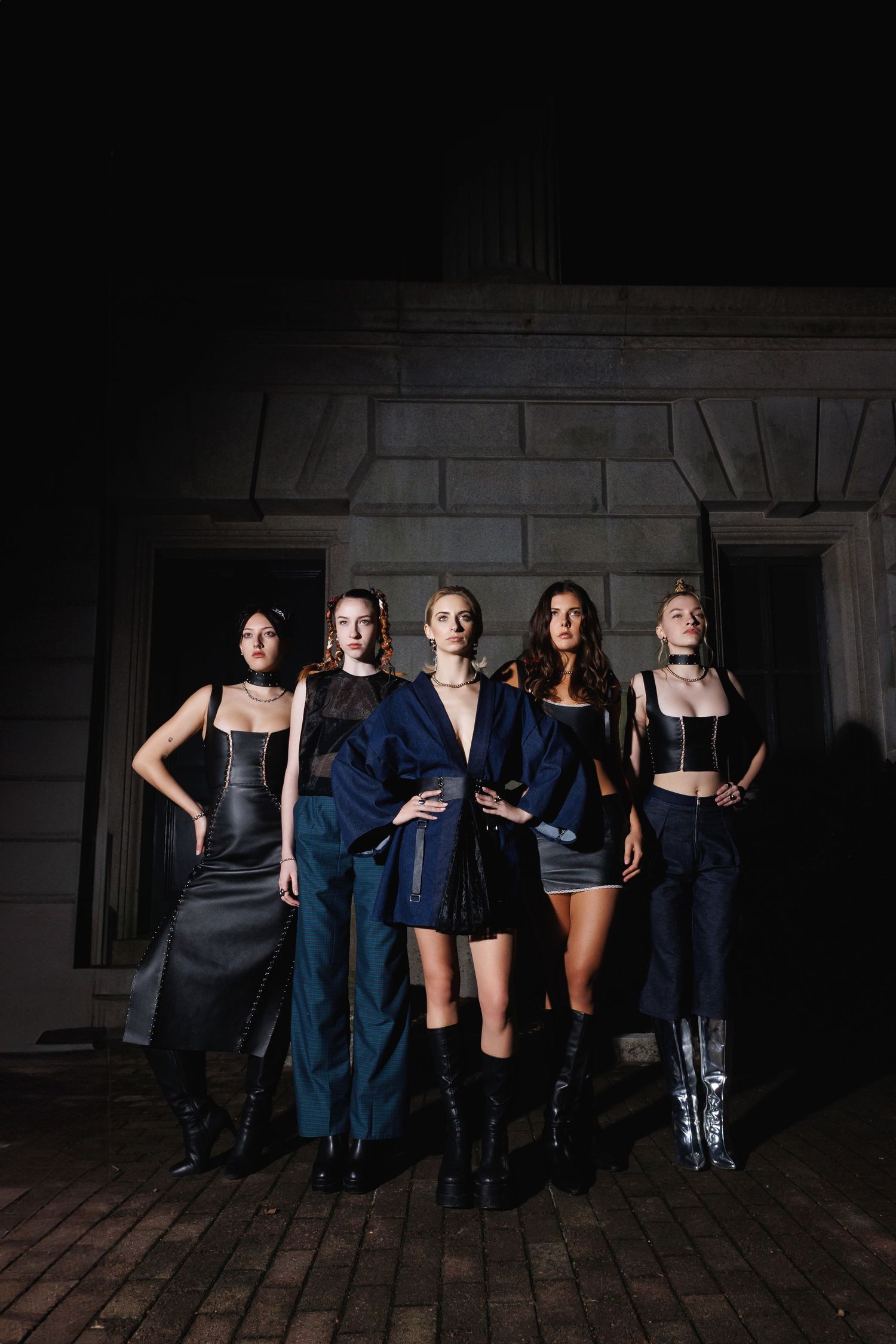A group poses together in dark outfits against a moody brick wall in an artistic photo series.