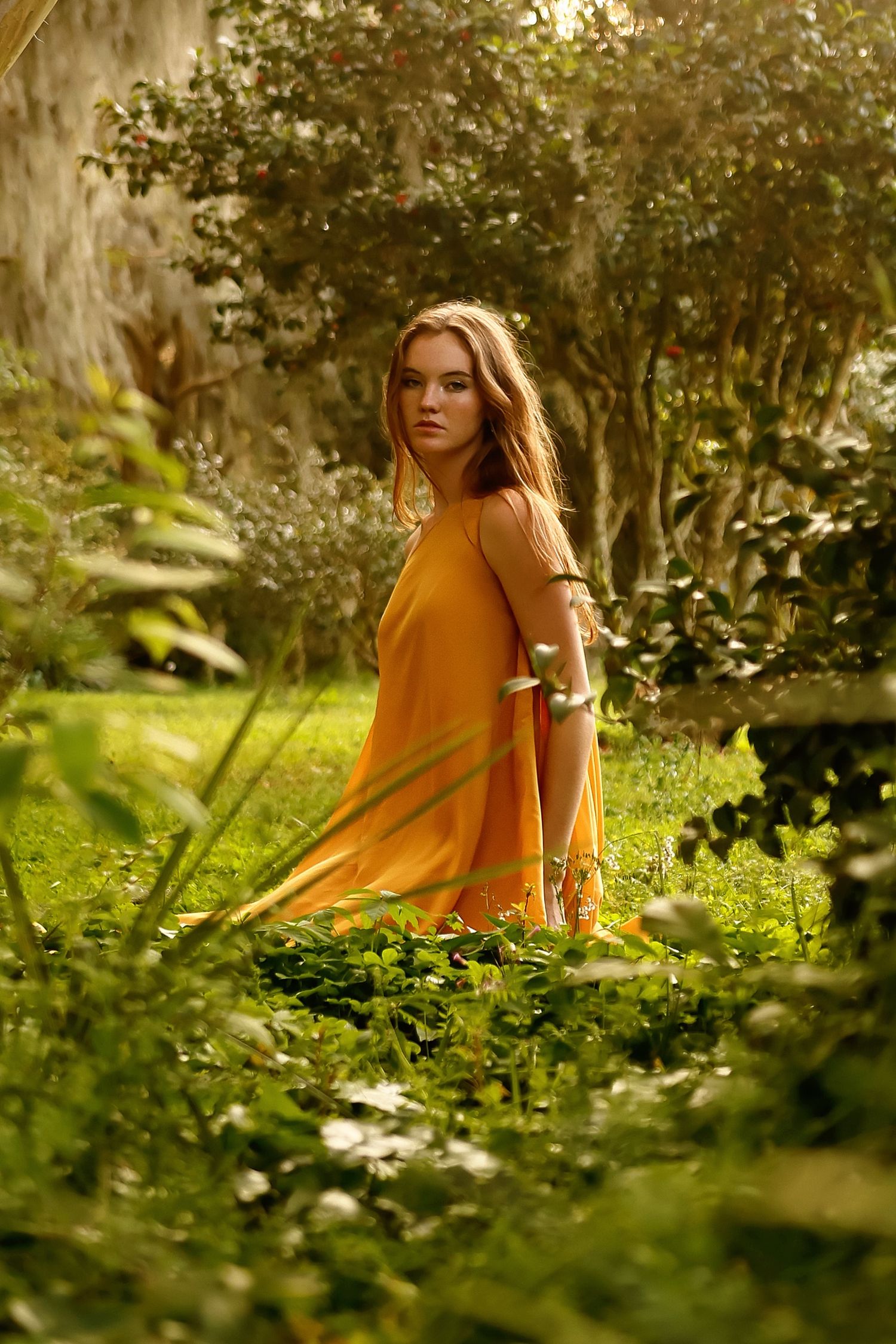 A person in a warm yellow sweater sits among lush green foliage in a sunlit garden setting.