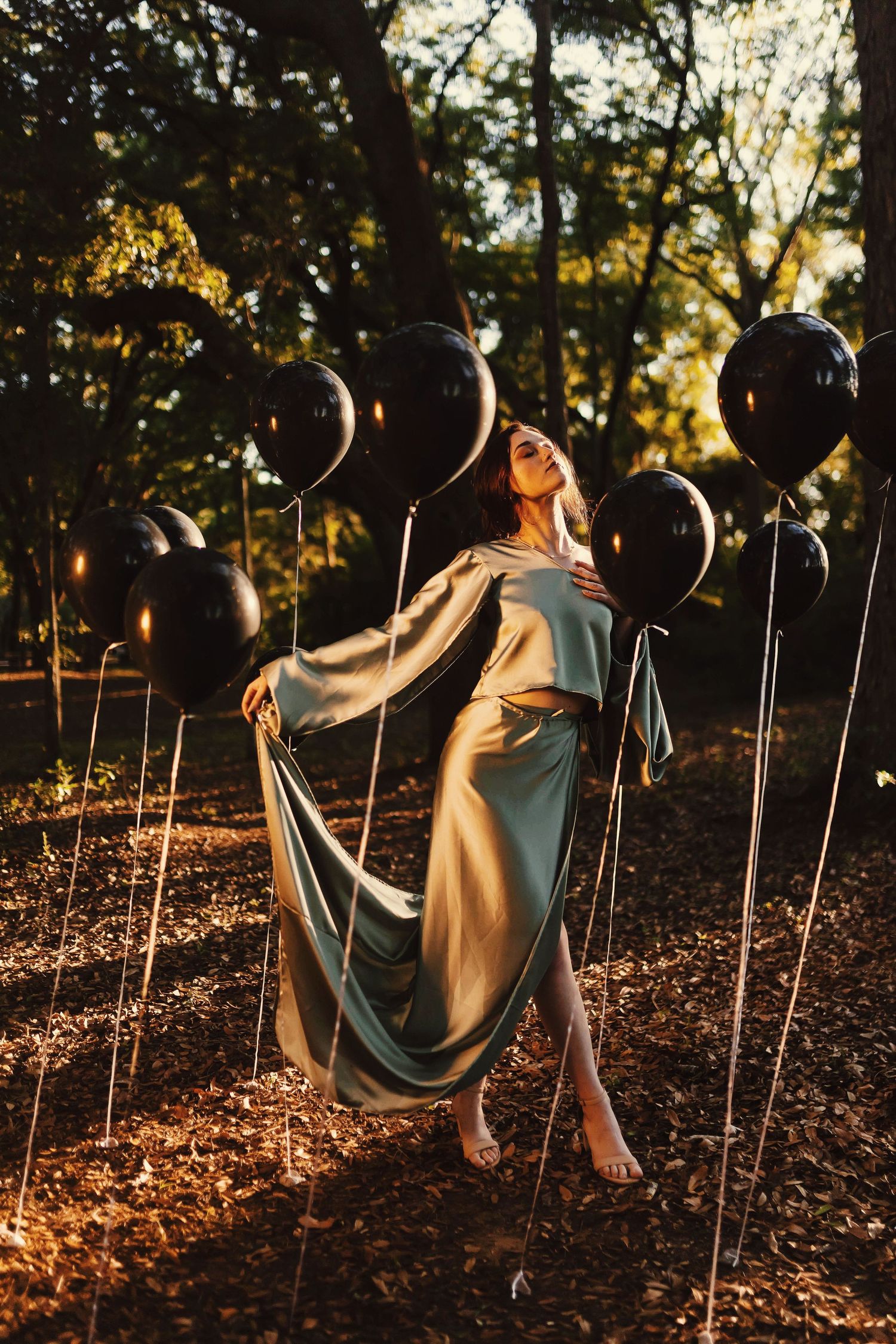 A person sits among black balloons in a wooded area during golden hour sunlight.