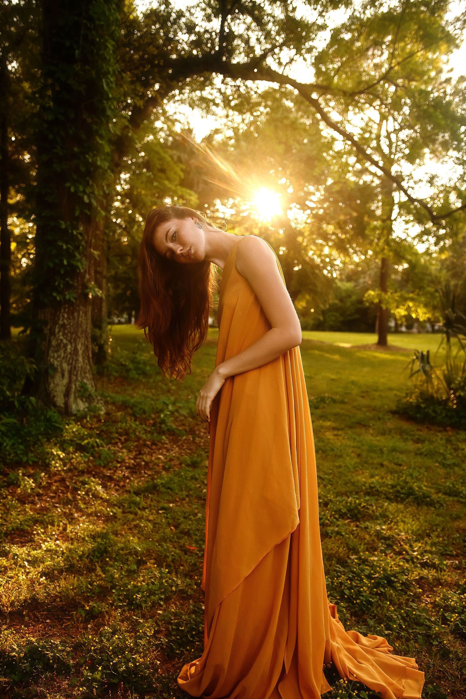 Person in flowing mustard yellow dress poses gracefully in a sunlit forest garden during golden hour.