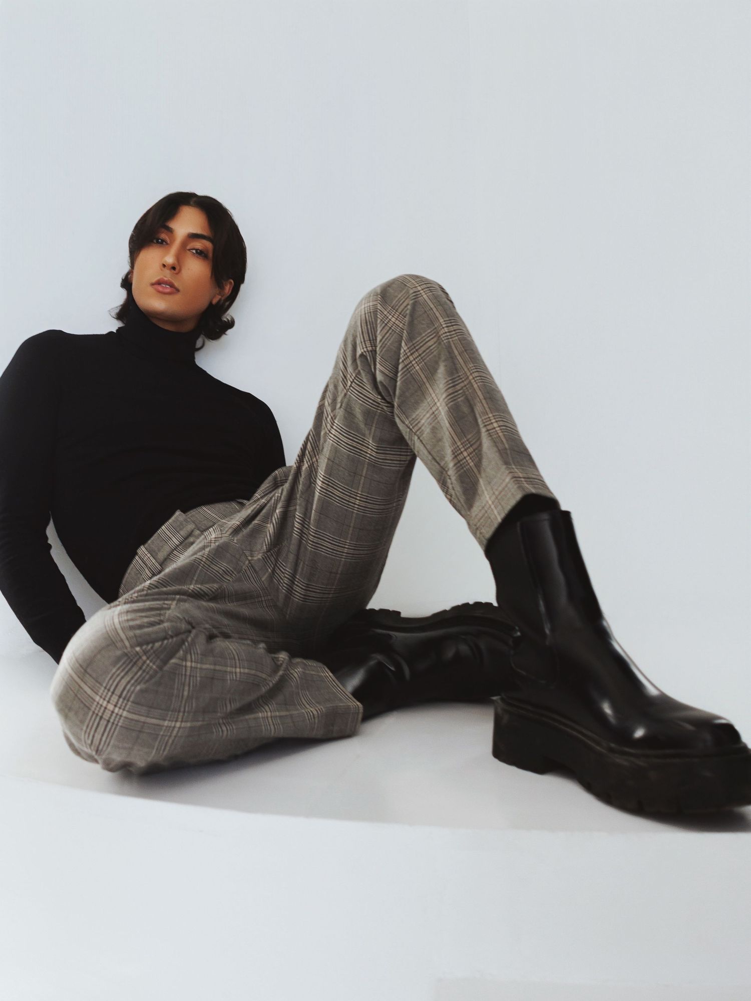 Fashion model in black turtleneck and plaid trousers poses with chunky black boots against white background.