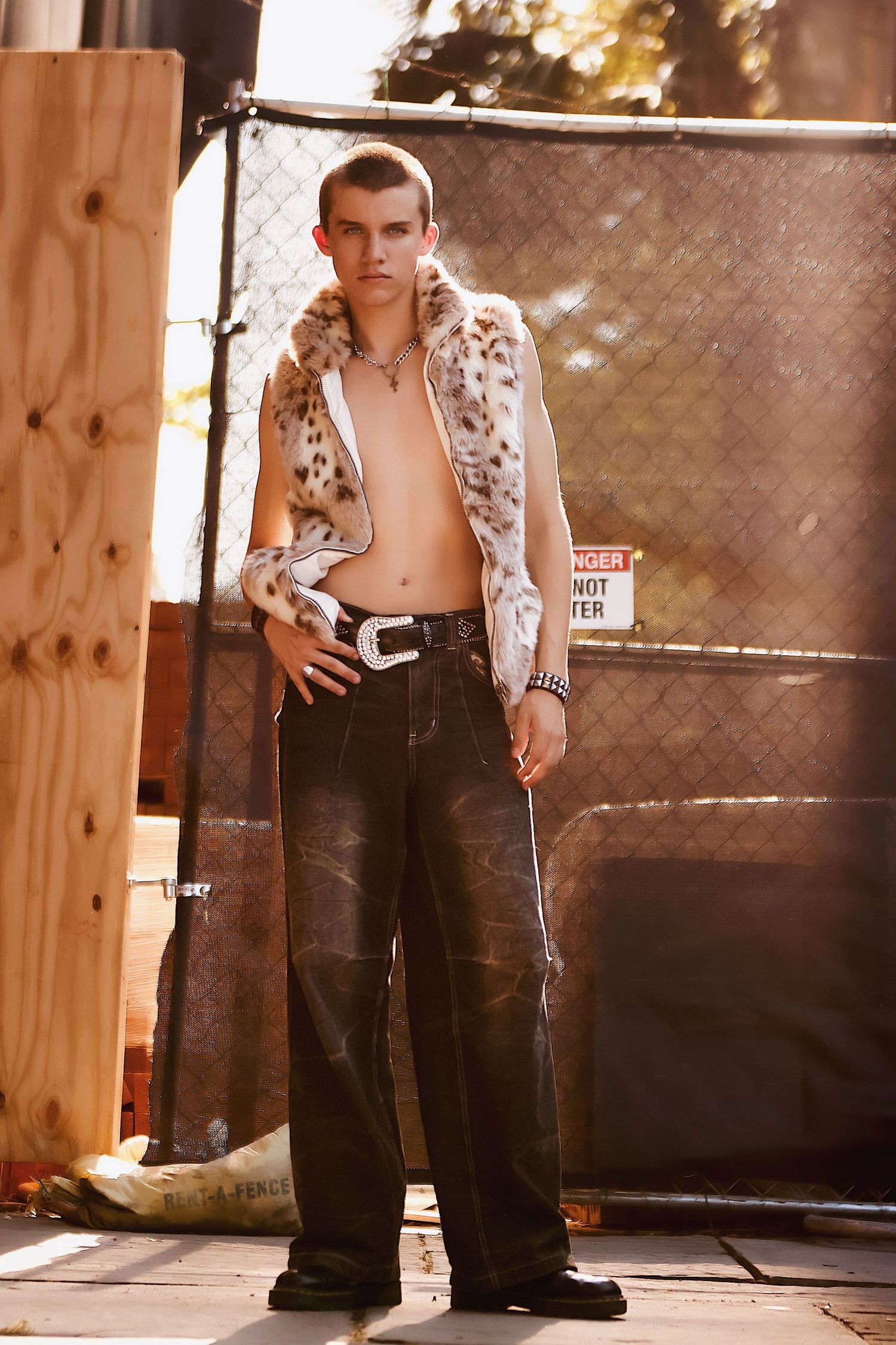 Fashion model poses outdoors wearing a spotted fur vest and dark wide-leg jeans against a chain-link fence in warm lighting.
