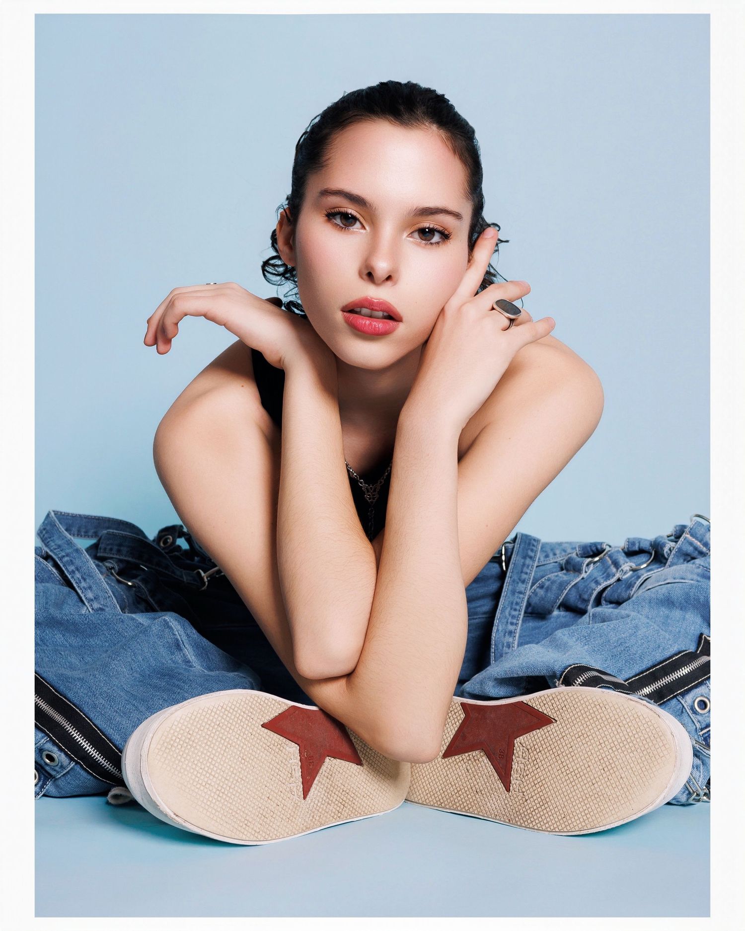 Fashion model in denim outfit sits cross-legged on blue backdrop wearing star-detailed shoes in editorial photoshoot.