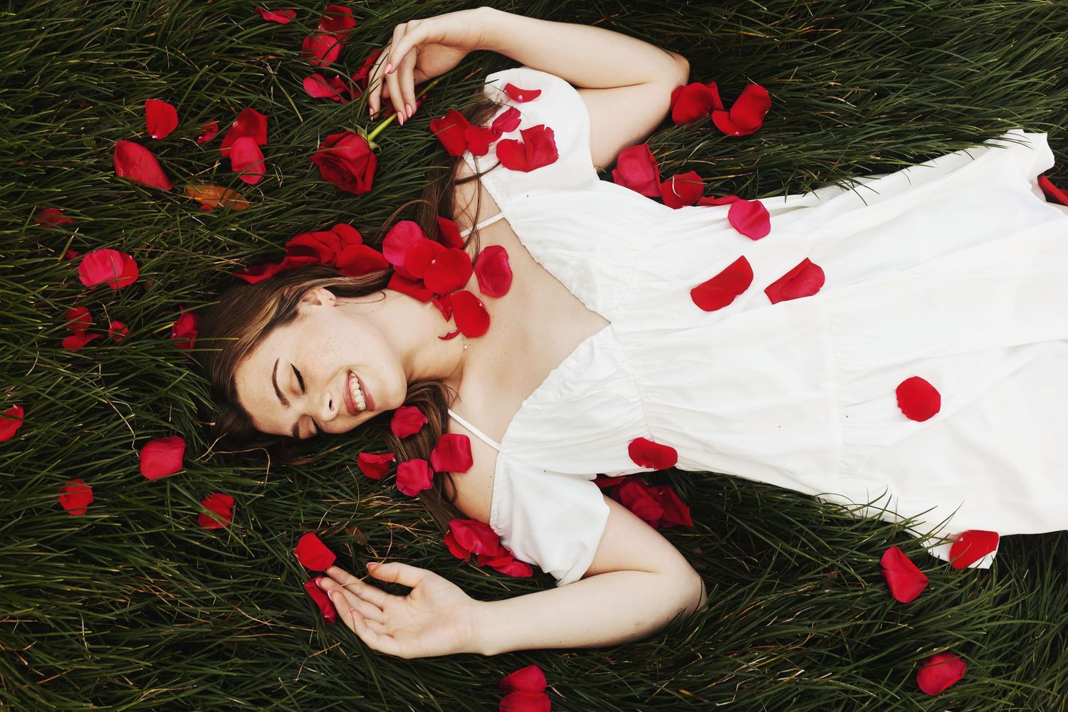 A figure in white lies on dark grass surrounded by scattered red rose petals in an artistic composition.