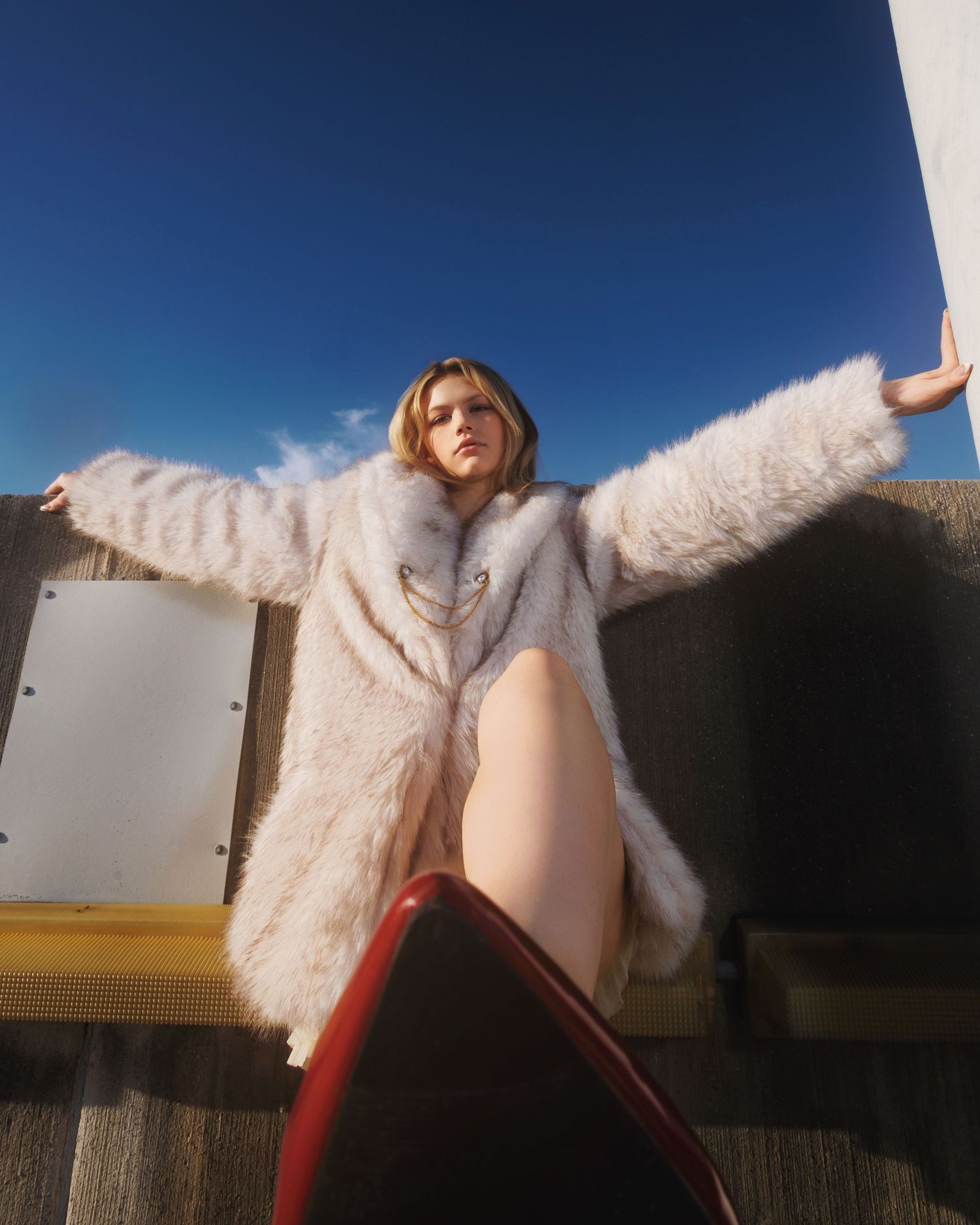A person in a fluffy white fur coat and red boots sits on a bench against a blue sky background.