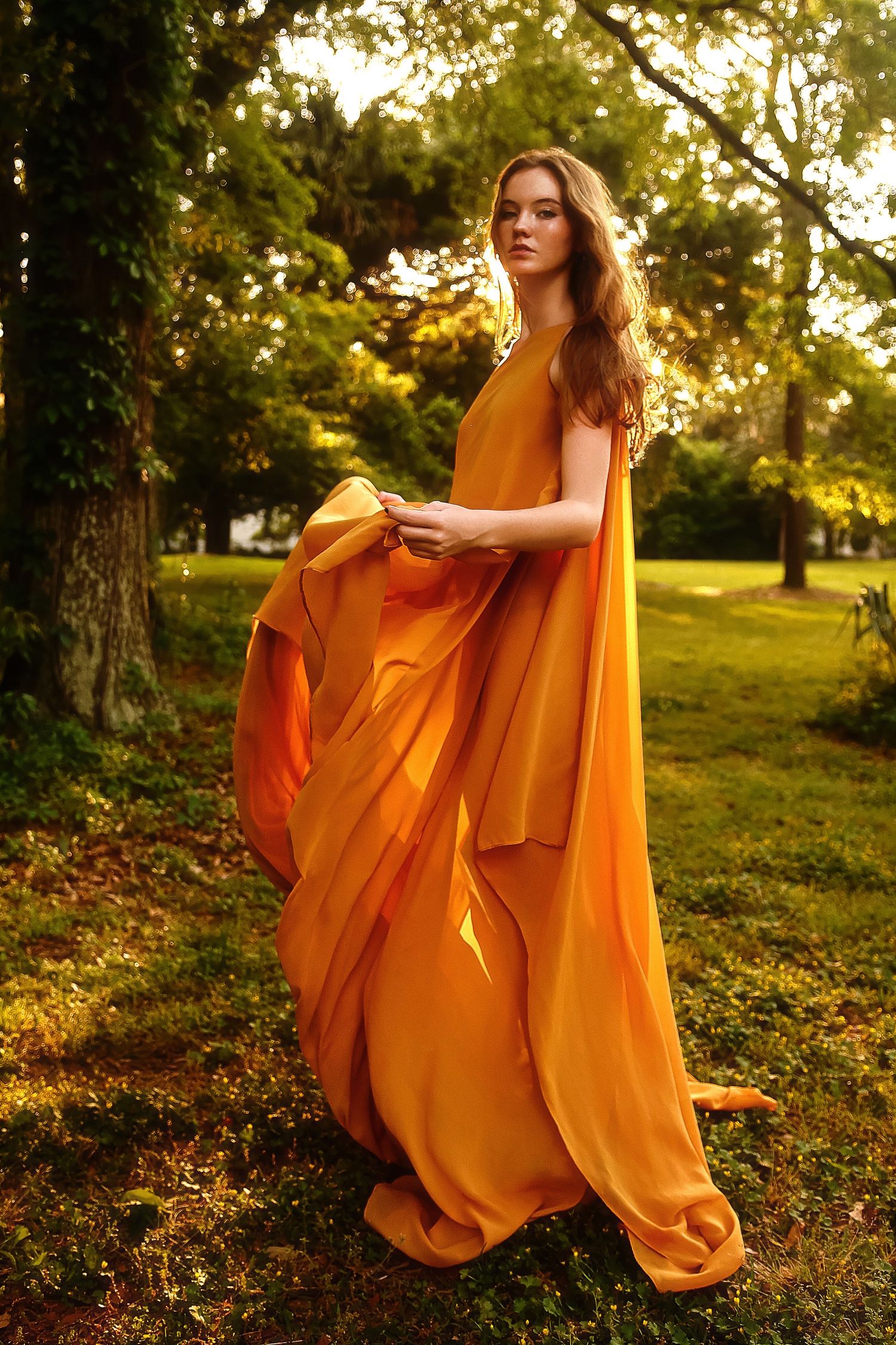 A flowing orange dress billows in the breeze as sunlight filters through autumn trees in a dreamy outdoor setting.