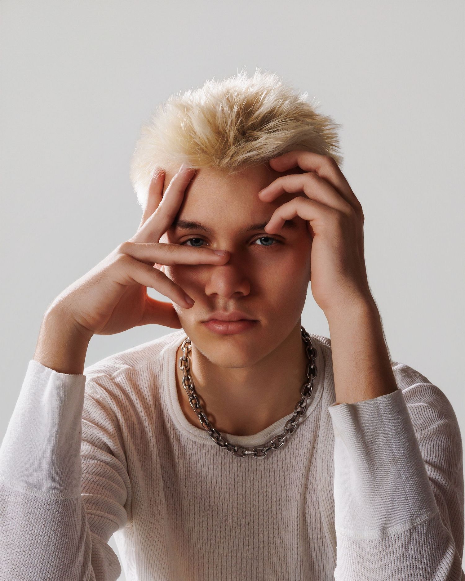Figure in white sweater and silver chain necklace poses with hands framing face against neutral background.