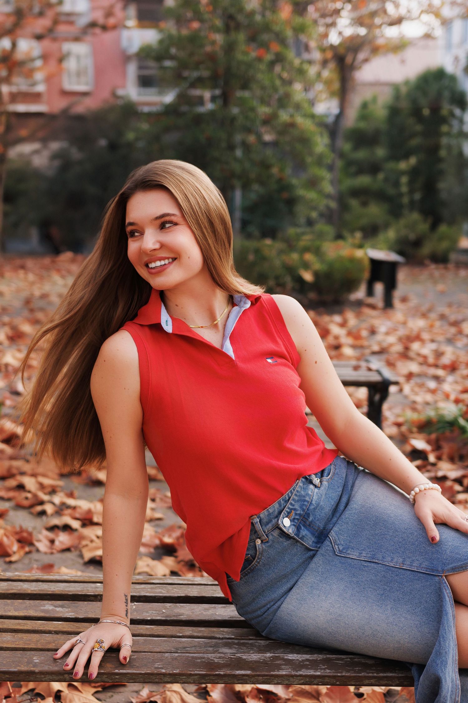 A person in a red sleeveless top and jeans sits on a bench surrounded by autumn leaves in a park.