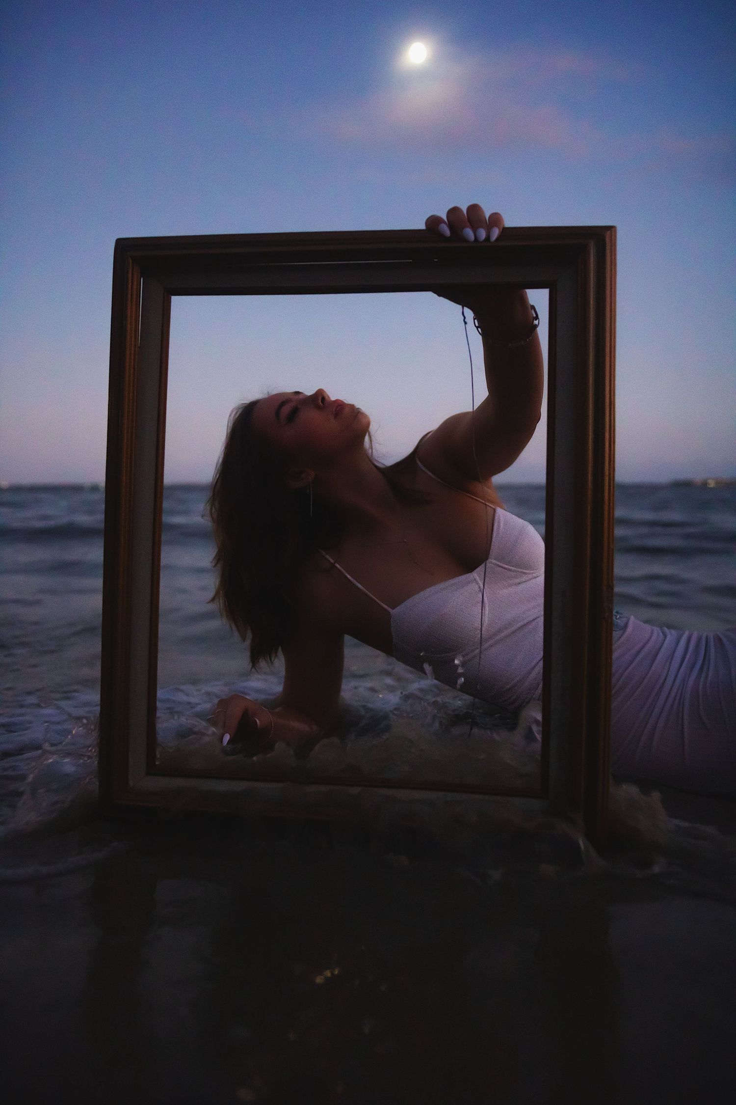 A silhouetted figure holds a picture frame against a dusky beach sky with full moon overhead.