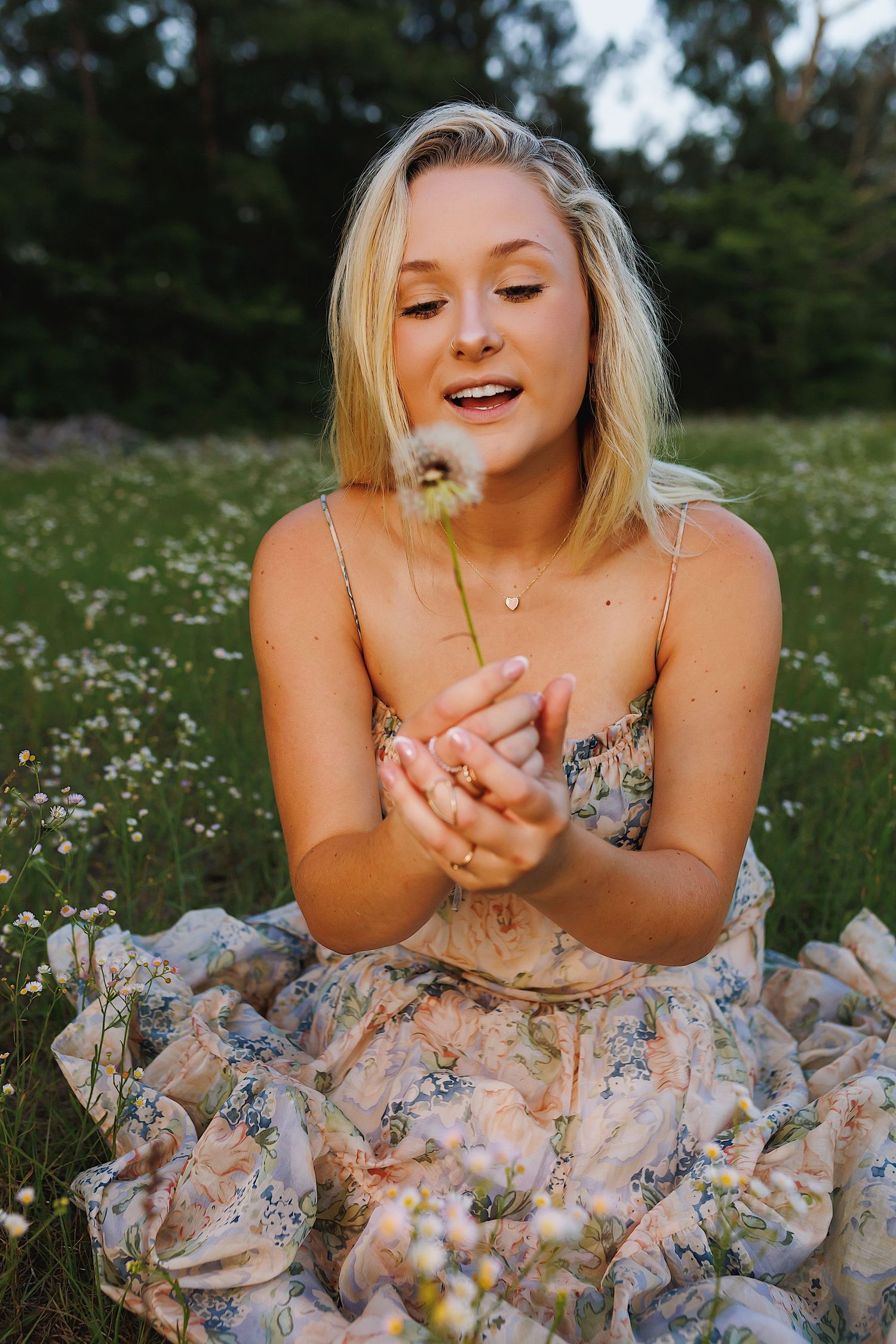 A person in a floral dress lies in a field of daisies while blowing flower petals from their hands.