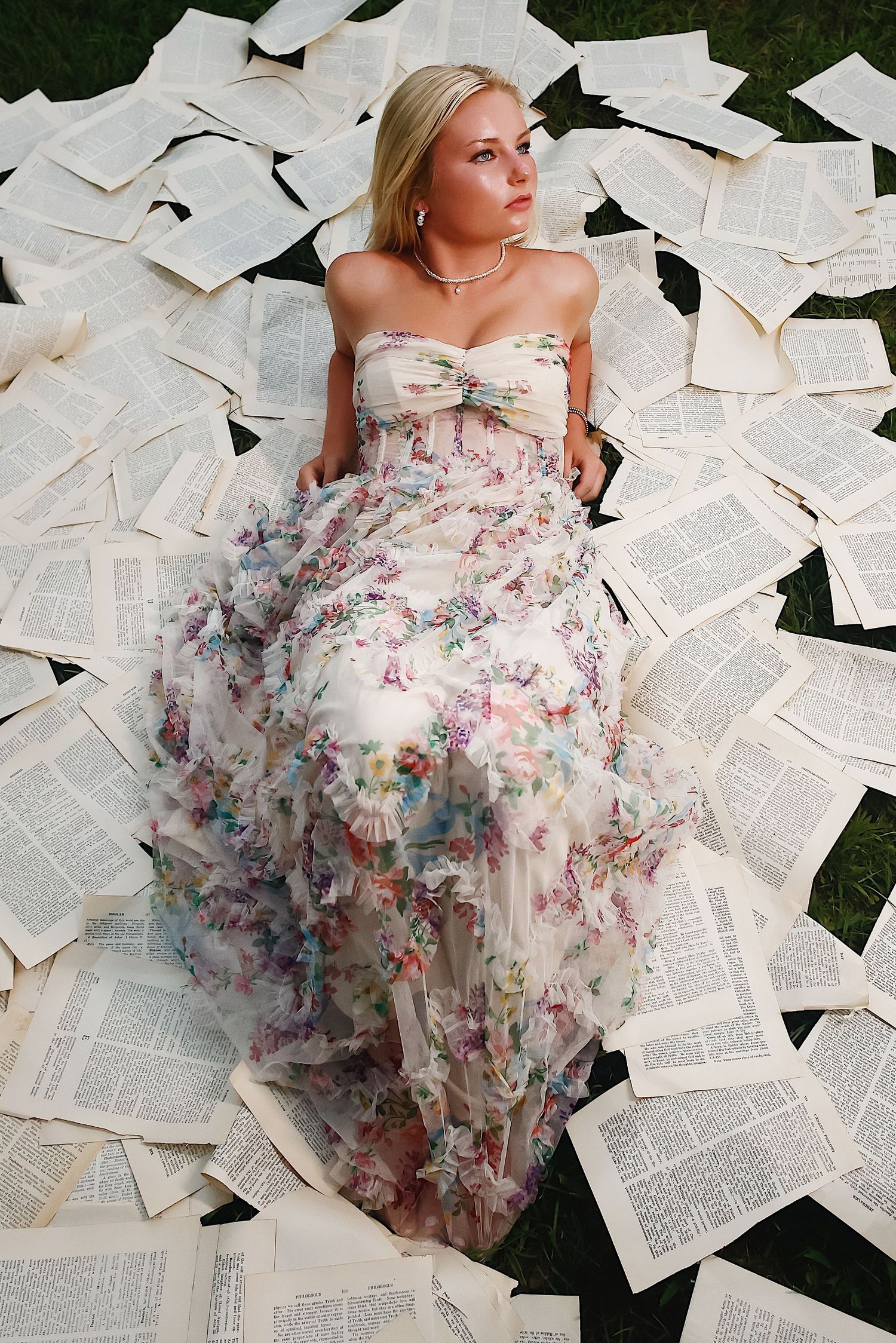 A flowing floral dress spreads across scattered book pages on grass in an artistic fashion photoshoot.