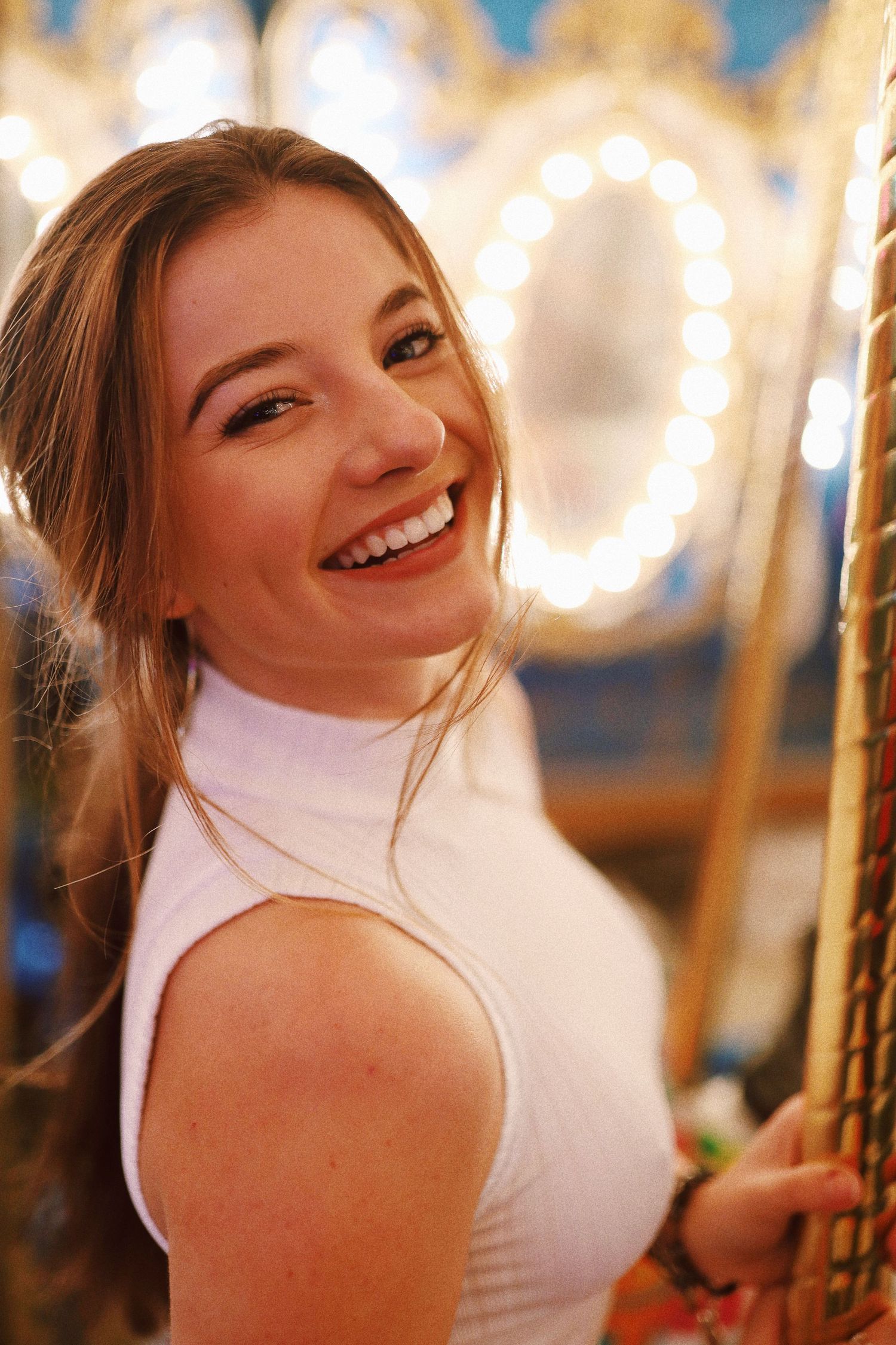 A person in a white turtleneck sweater smiles warmly while standing near glowing carousel lights.
