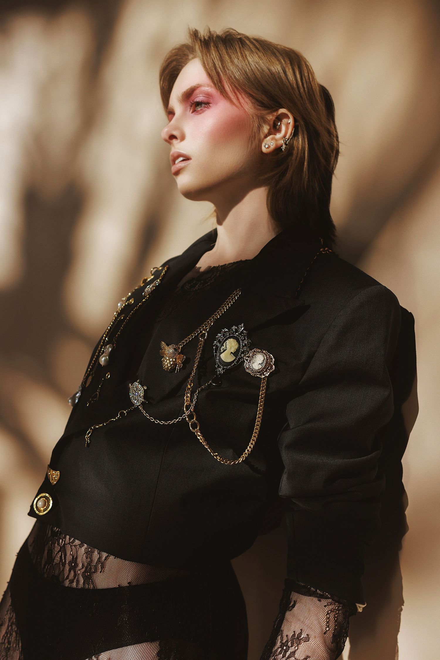Fashion model in black lace and military-style jacket adorned with vintage brooches and chains poses in dramatic lighting.