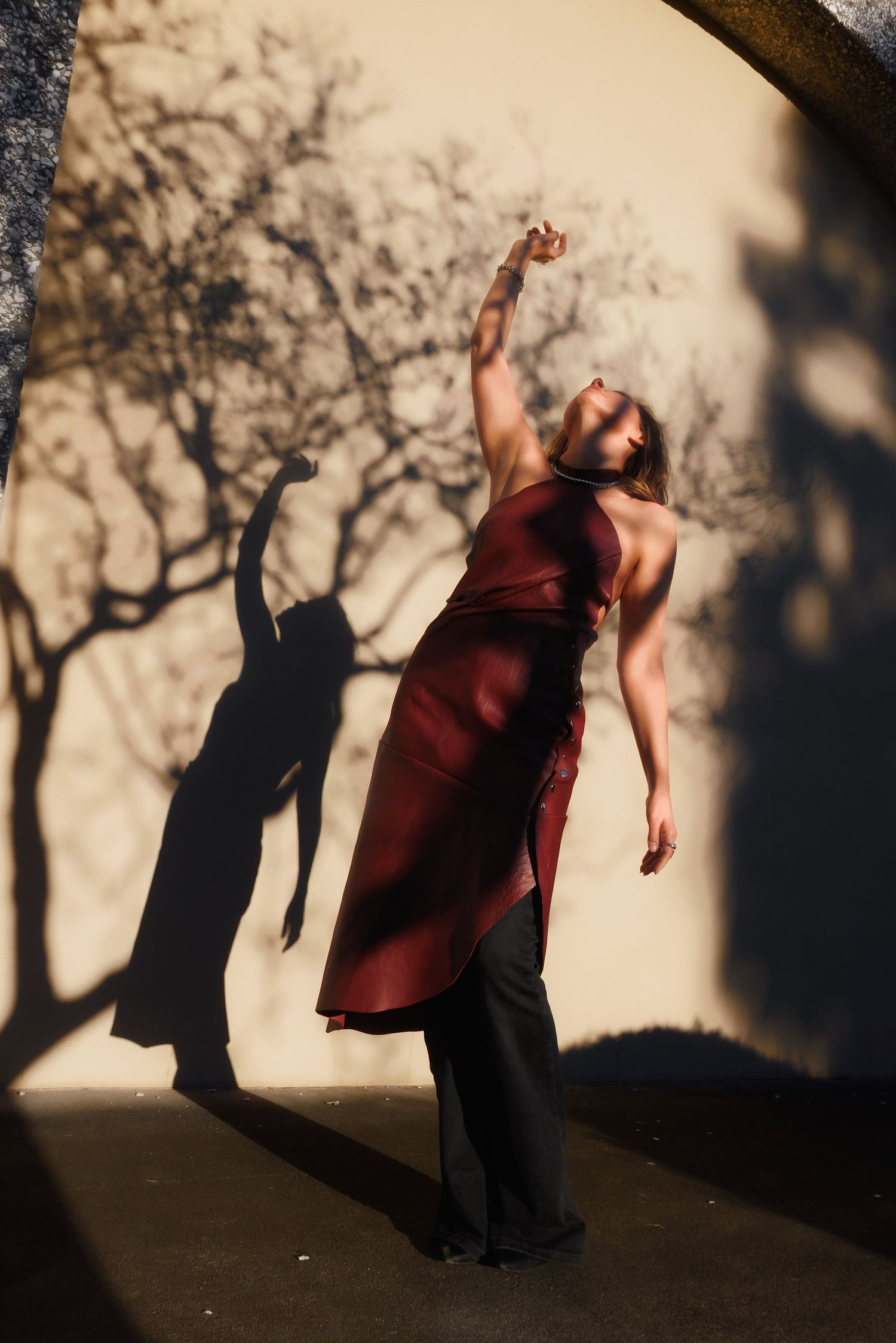 A figure performs an expressive dance movement against a shadow-cast wall with tree silhouettes in reddish lighting.