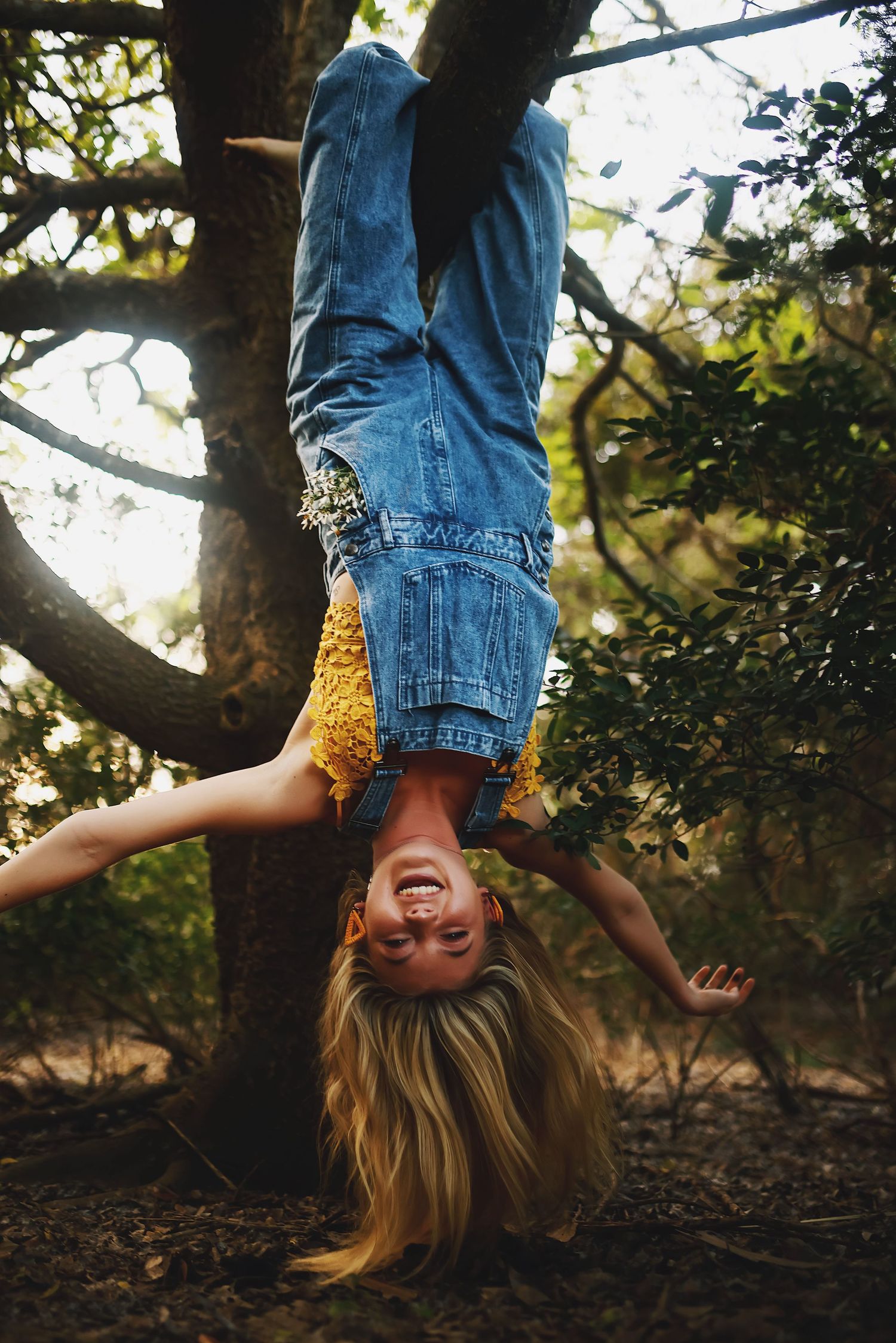A person in denim jeans hangs upside down from a tree branch during golden hour lighting.