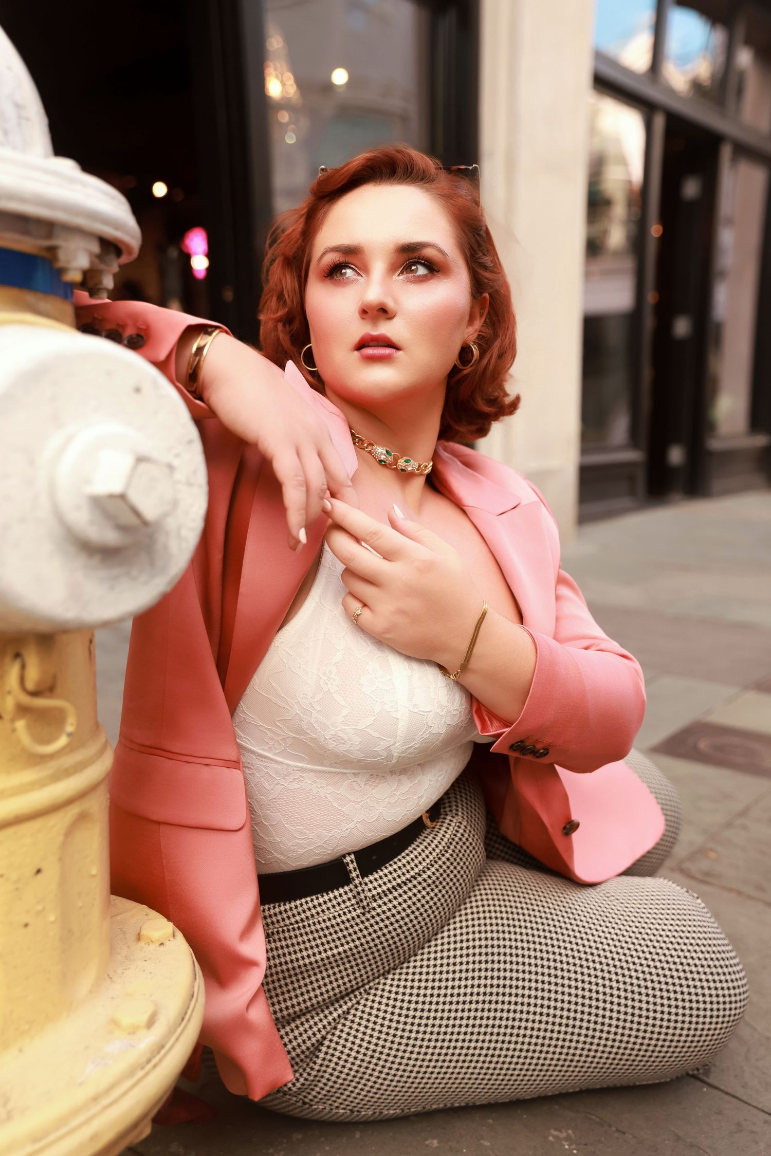 Fashion model in pink blazer and houndstooth pants poses elegantly against vintage fire hydrant on city street.