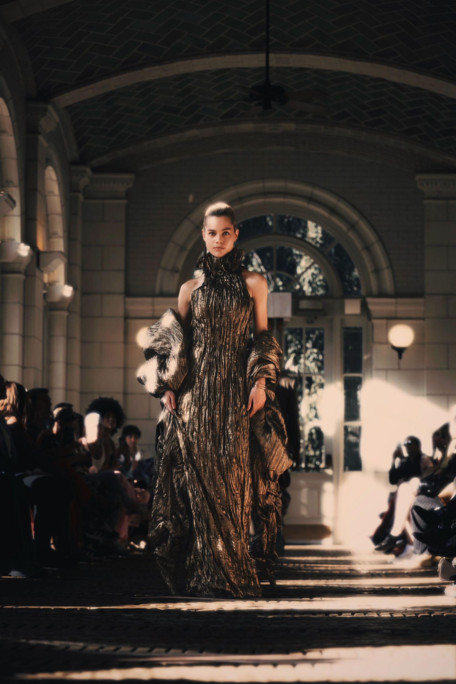 A fashion show model walks down a dark runway wearing an avant-garde dress made of textured natural materials.