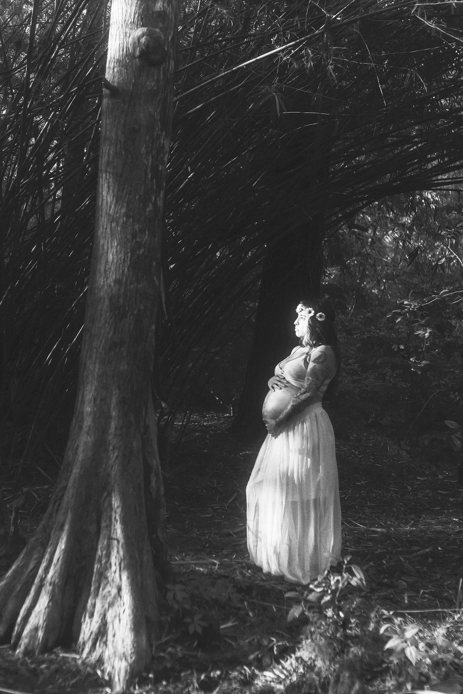 Black and white photo of pregnant woman in white dress standing among trees in a forest, cradling her baby bump.