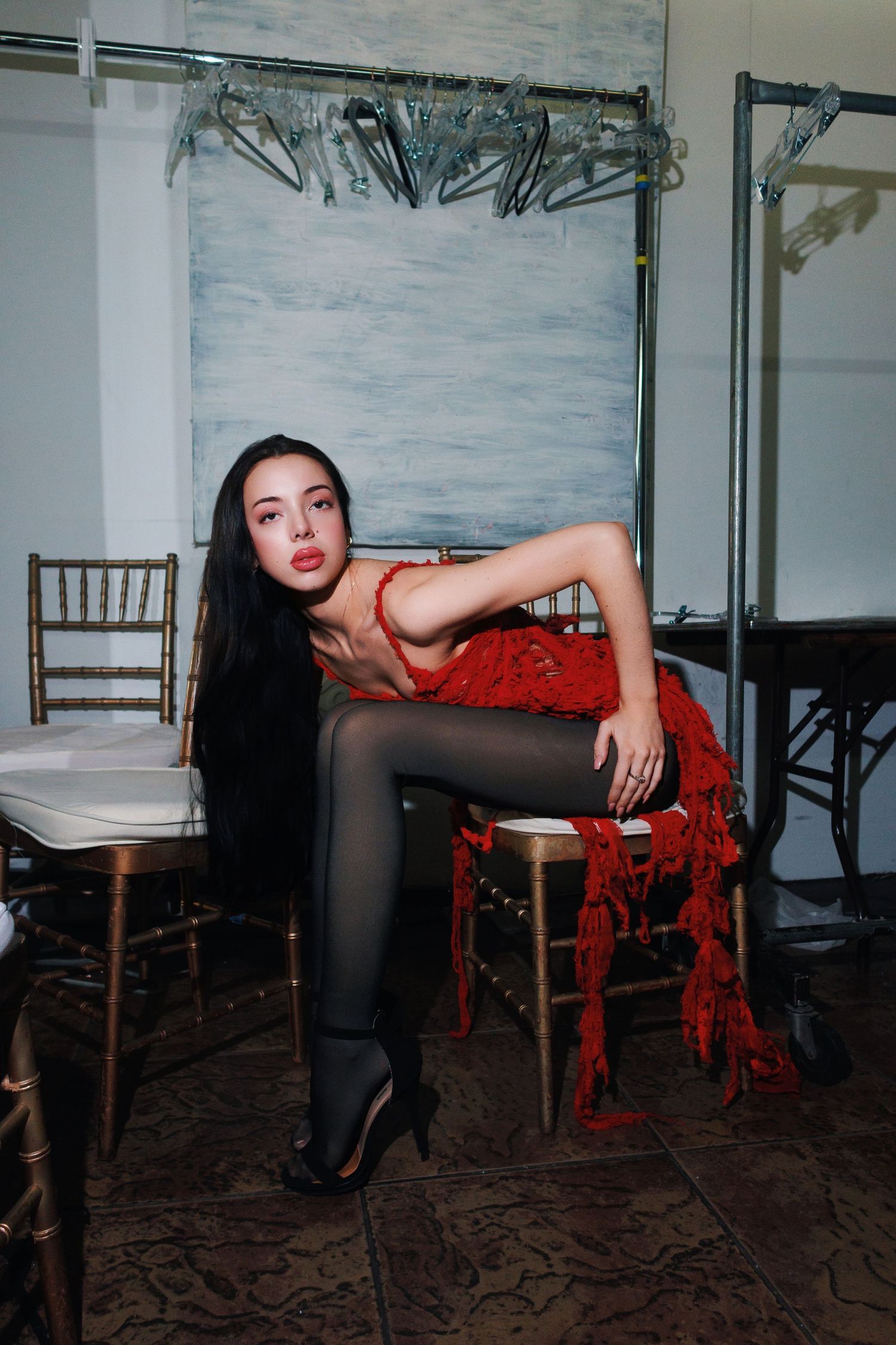 Artistic portrait of a person in a red fringe dress and black tights posing on a chair in a minimalist room setting.