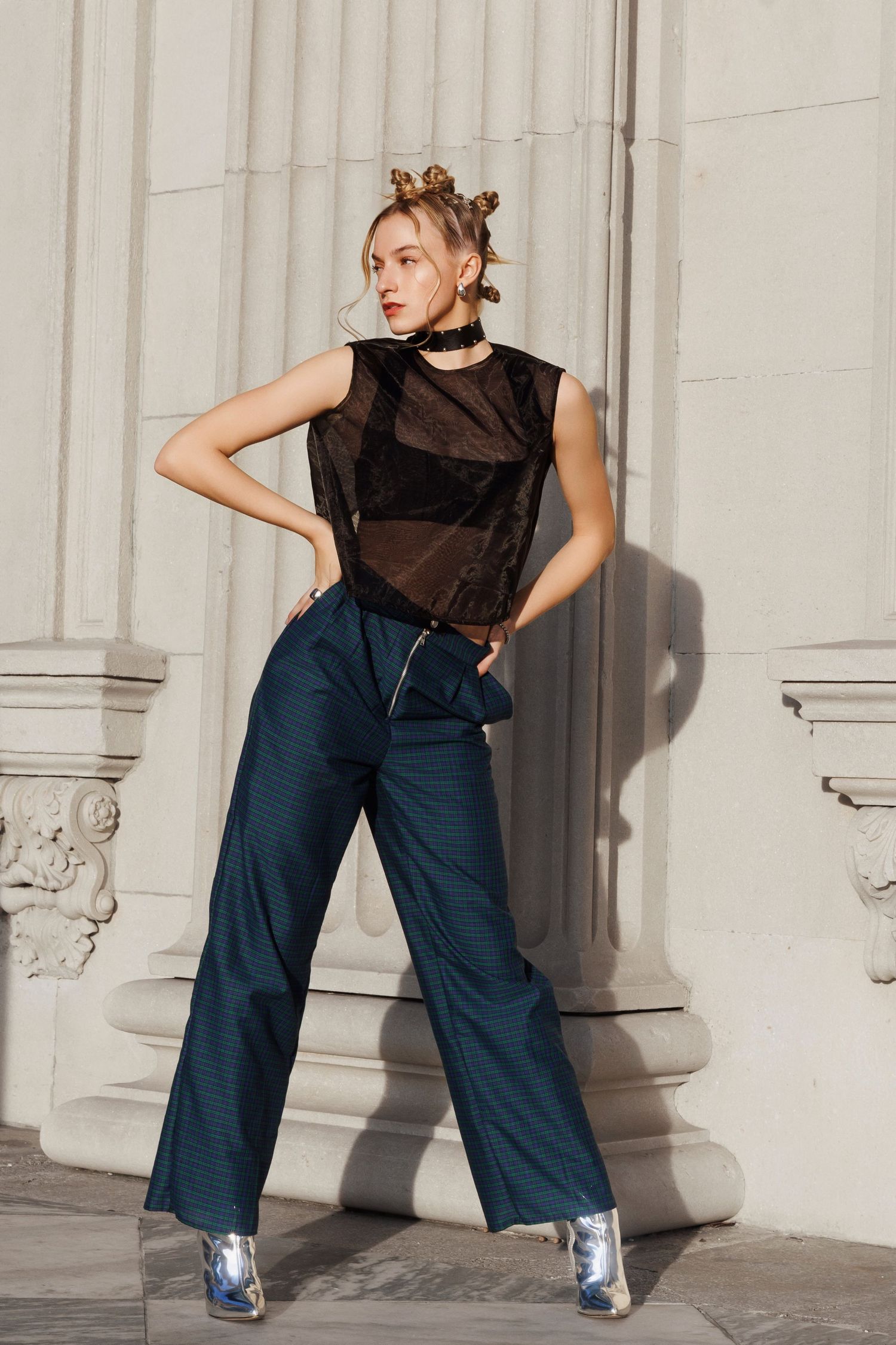 Fashion model in a sheer black sleeveless top, wide-leg navy pants and metallic boots poses against white architectural columns.